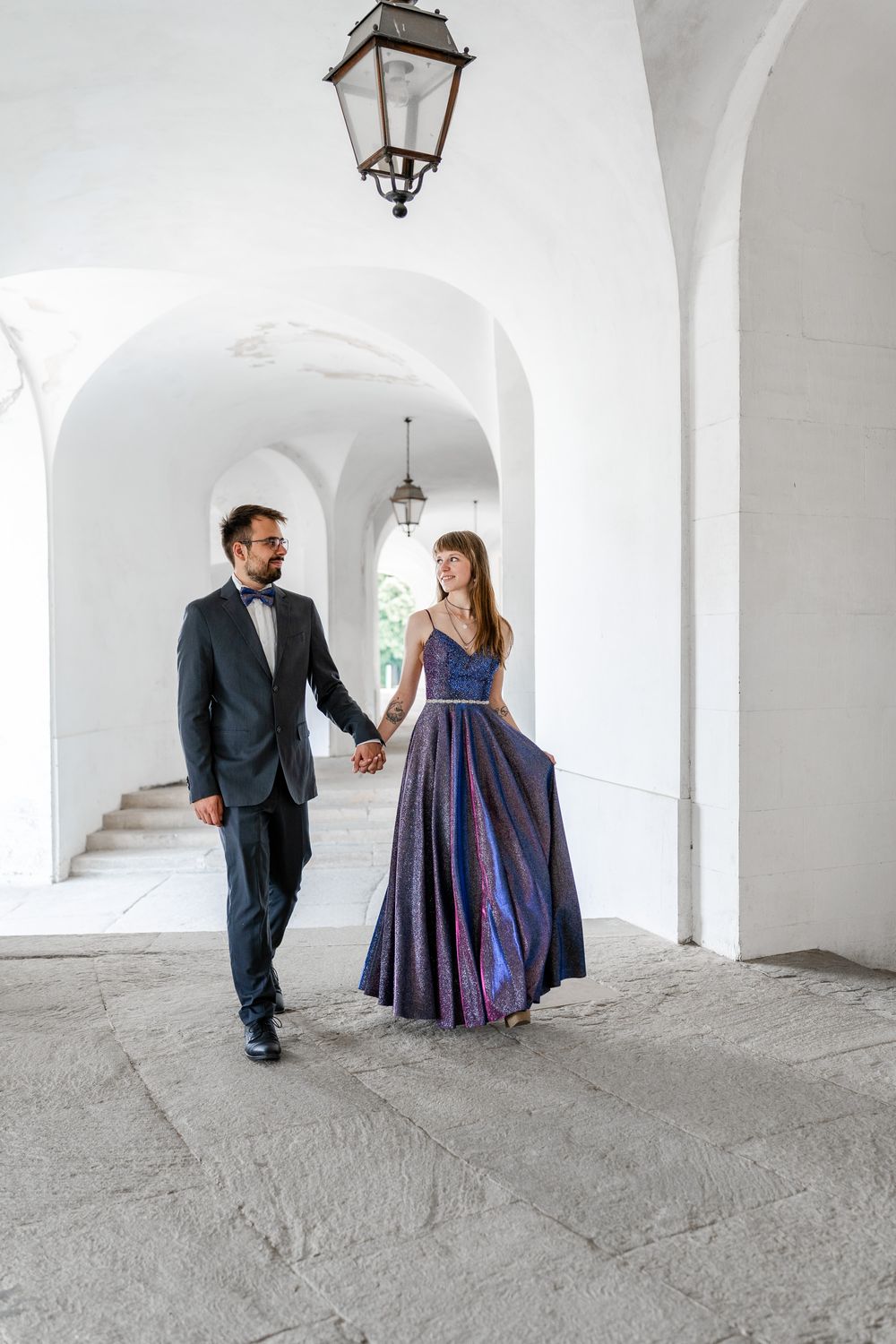 Elegant couple walking through white arched corridor with hanging lanterns.