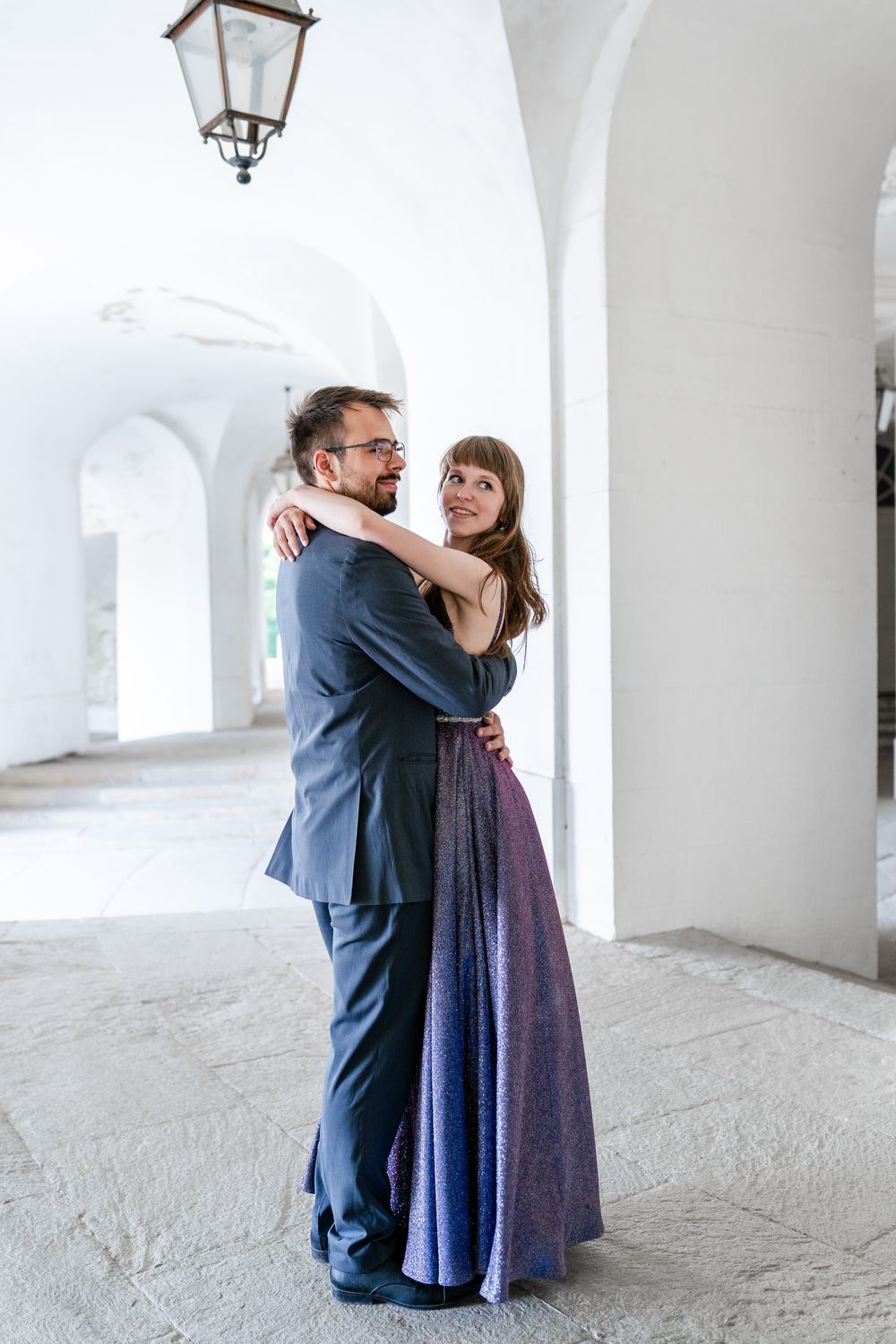 Couple embracing under white arched hallway with vintage lanterns overhead.