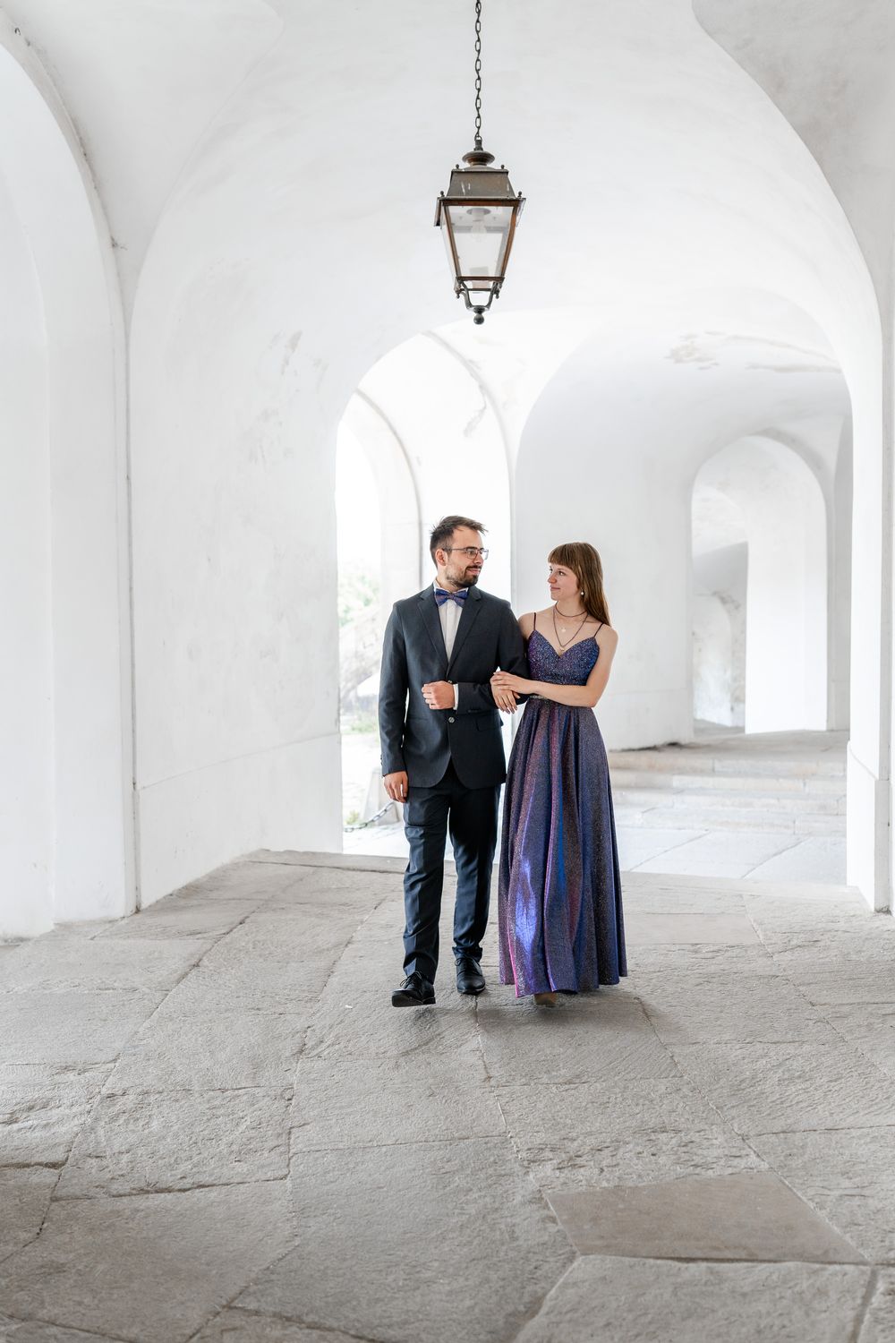 Couple in formal attire walks through an elegant white archway with hanging lantern.