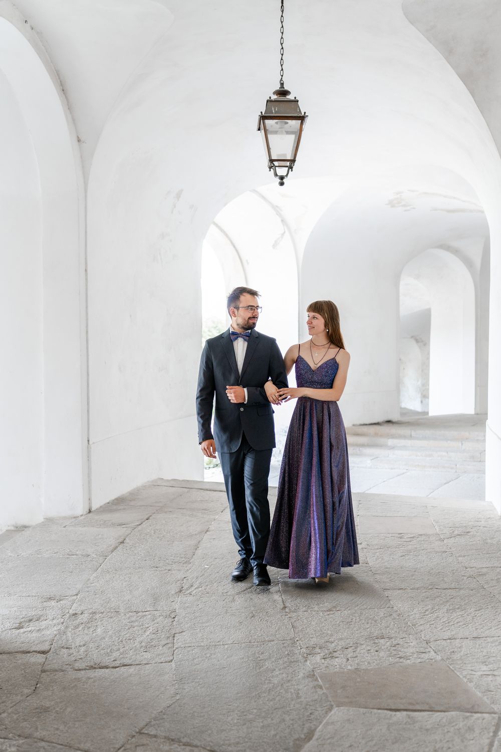 Formal couple poses in ornate white hallway beneath antique lantern.