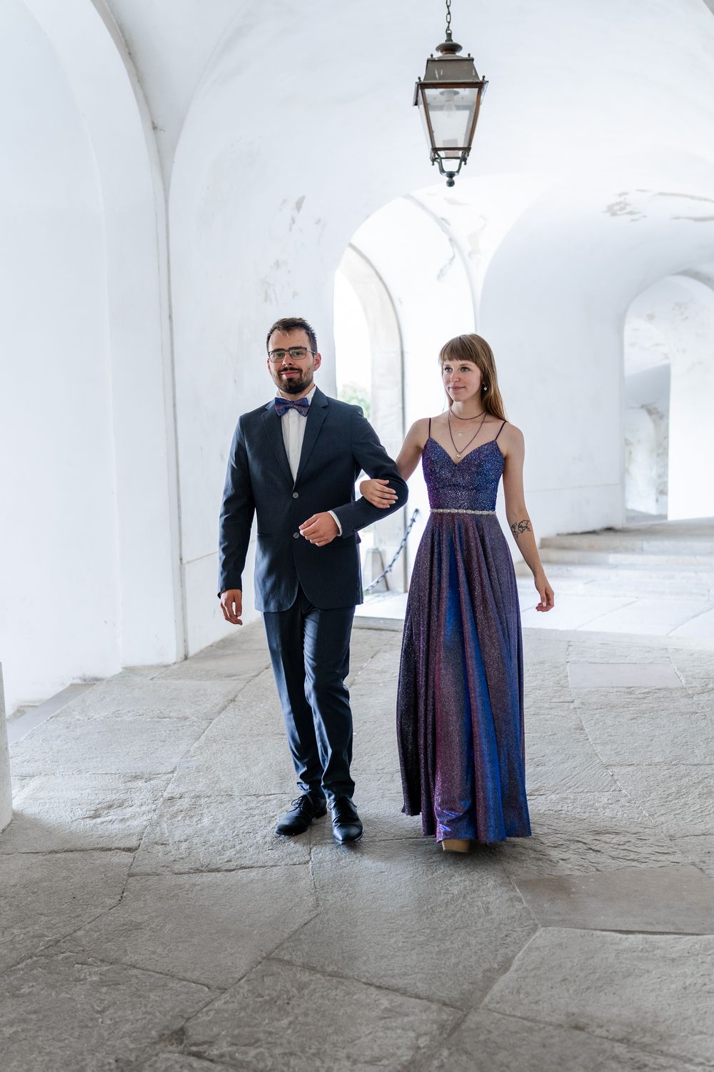 Elegant couple strolling through white arched corridor with vintage lighting.