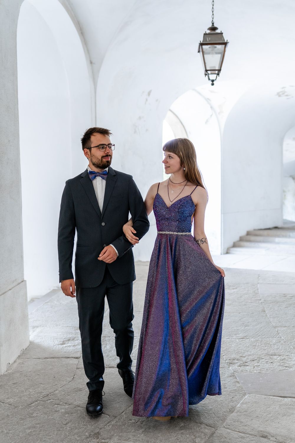 Couple in evening wear walks through bright arched passageway with vintage lighting.