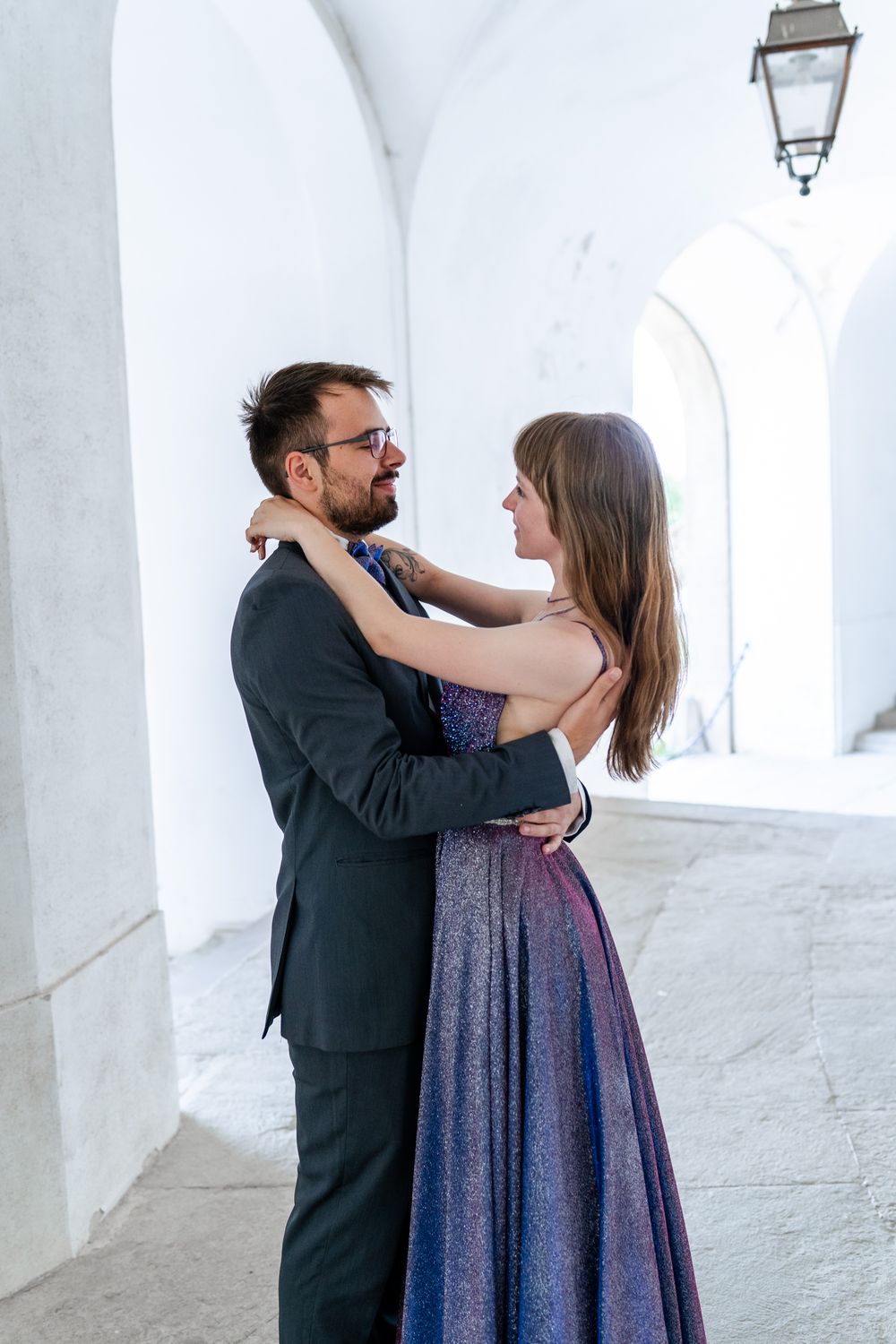 Romantic embrace in formal attire under white arched hallway with hanging lamp.