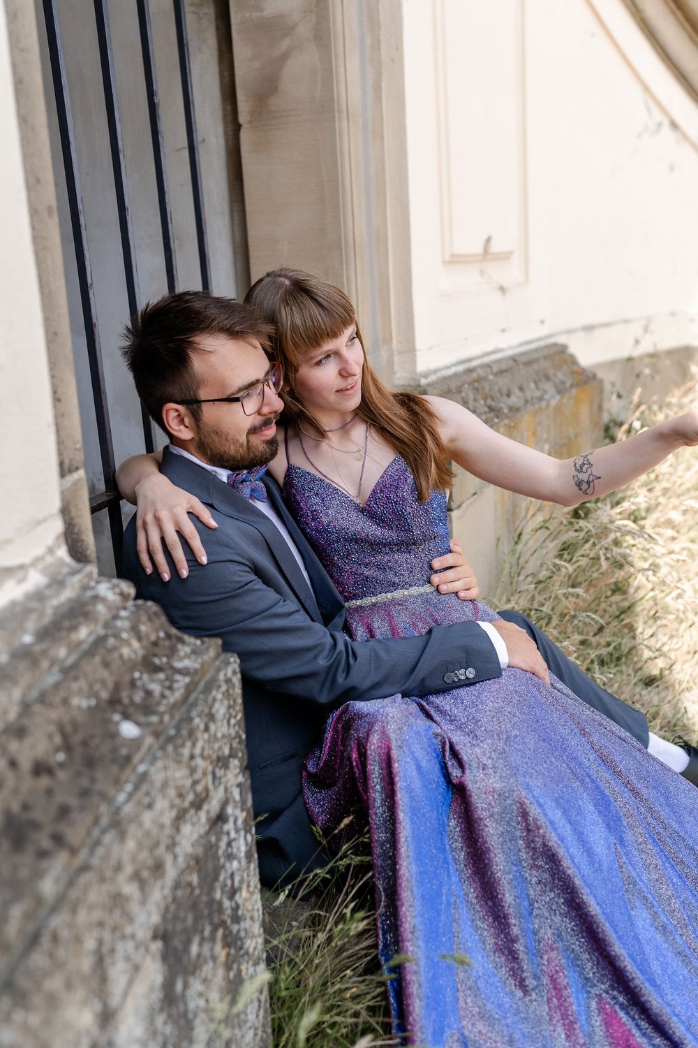 Couple in formal attire sitting by stone wall and iron gate.