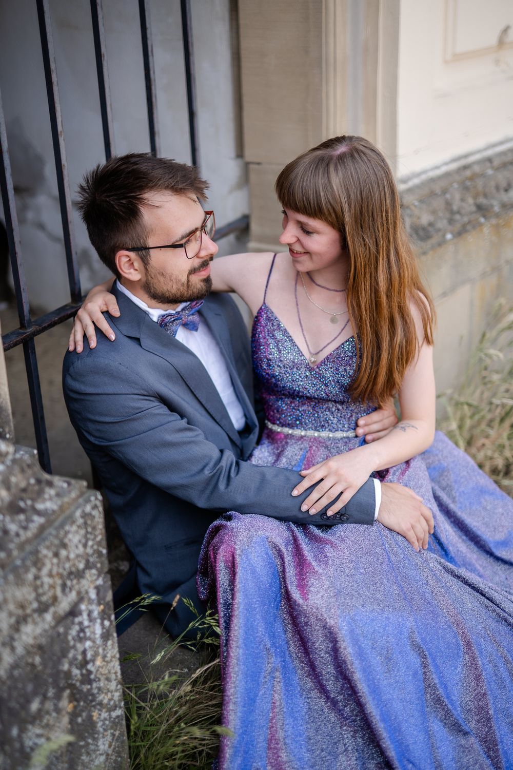Couple shares intimate moment while sitting by iron gate in formal wear.