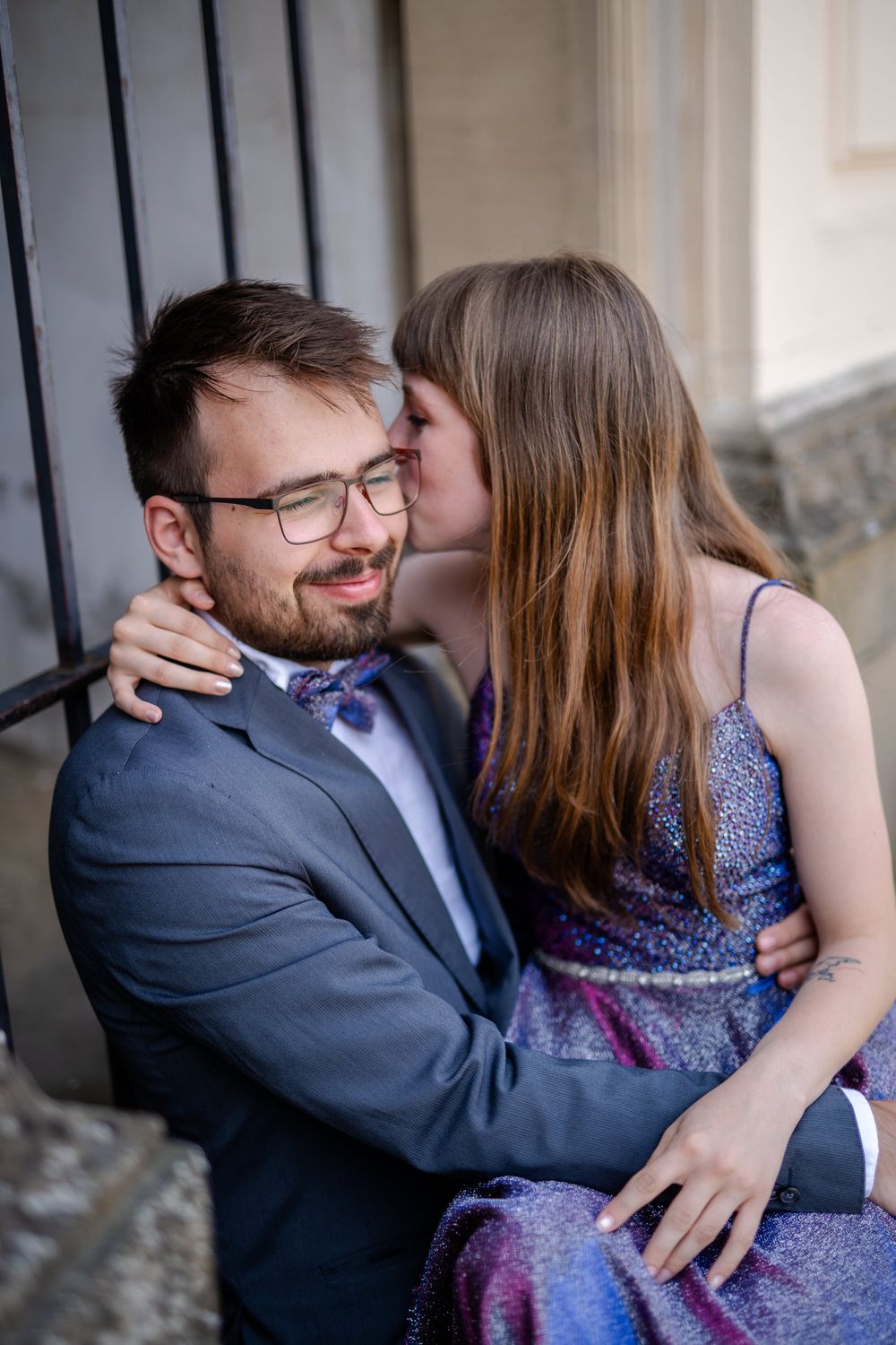 Romantic couple embraces against stone wall, with her in a sparkly purple dress and him in a navy suit.