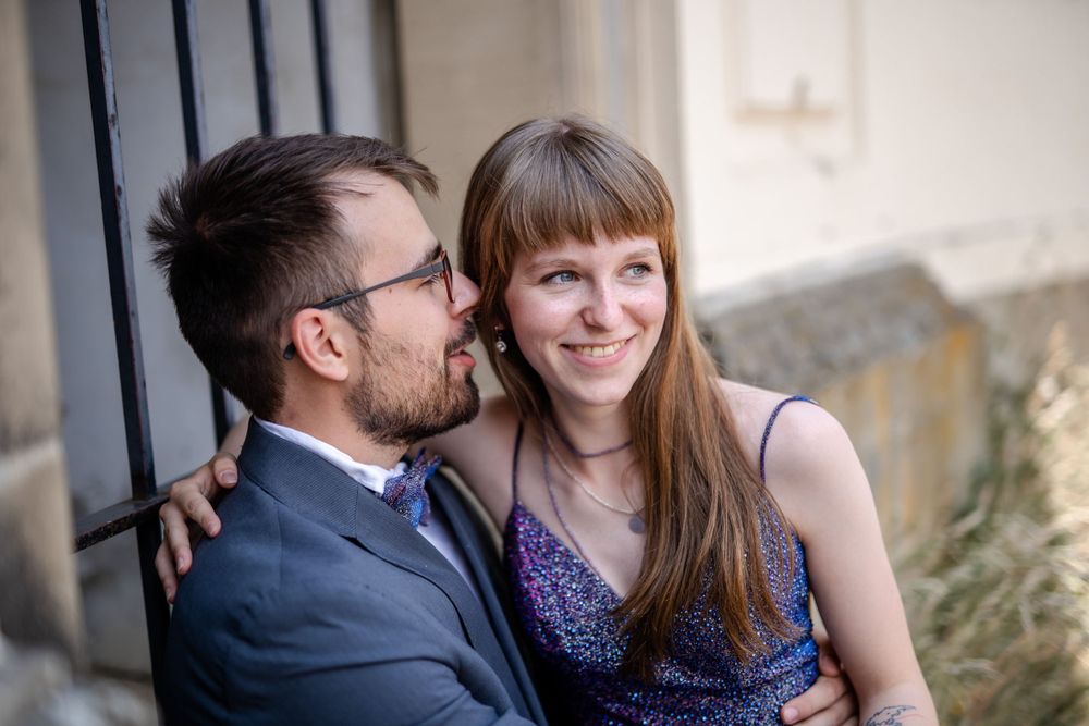 Couple in formal attire smile while sitting by stone wall.