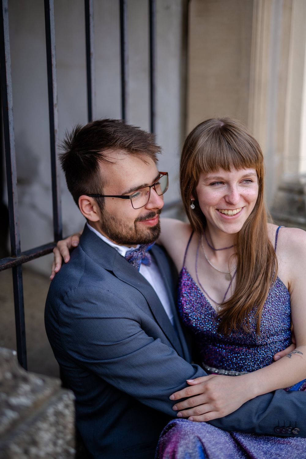 Happy couple snuggling together while seated, wearing formal attire near vintage architecture.