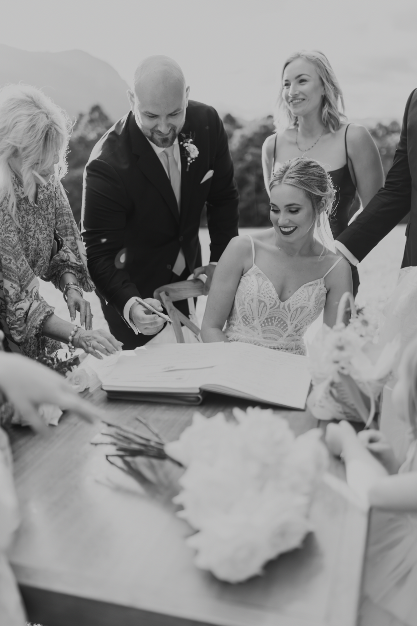 A young bride sits surrounded by her husband, maid of honour and celebrant as the groom signs the marriage certificate.