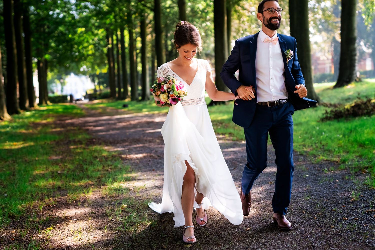 Couple walking together on a tree-lined path in wedding attire.