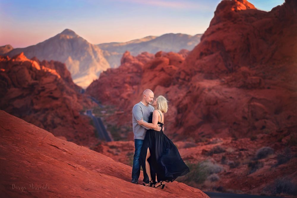 Couple embracing on red rocks with dramatic mountain vista in background during sunset.