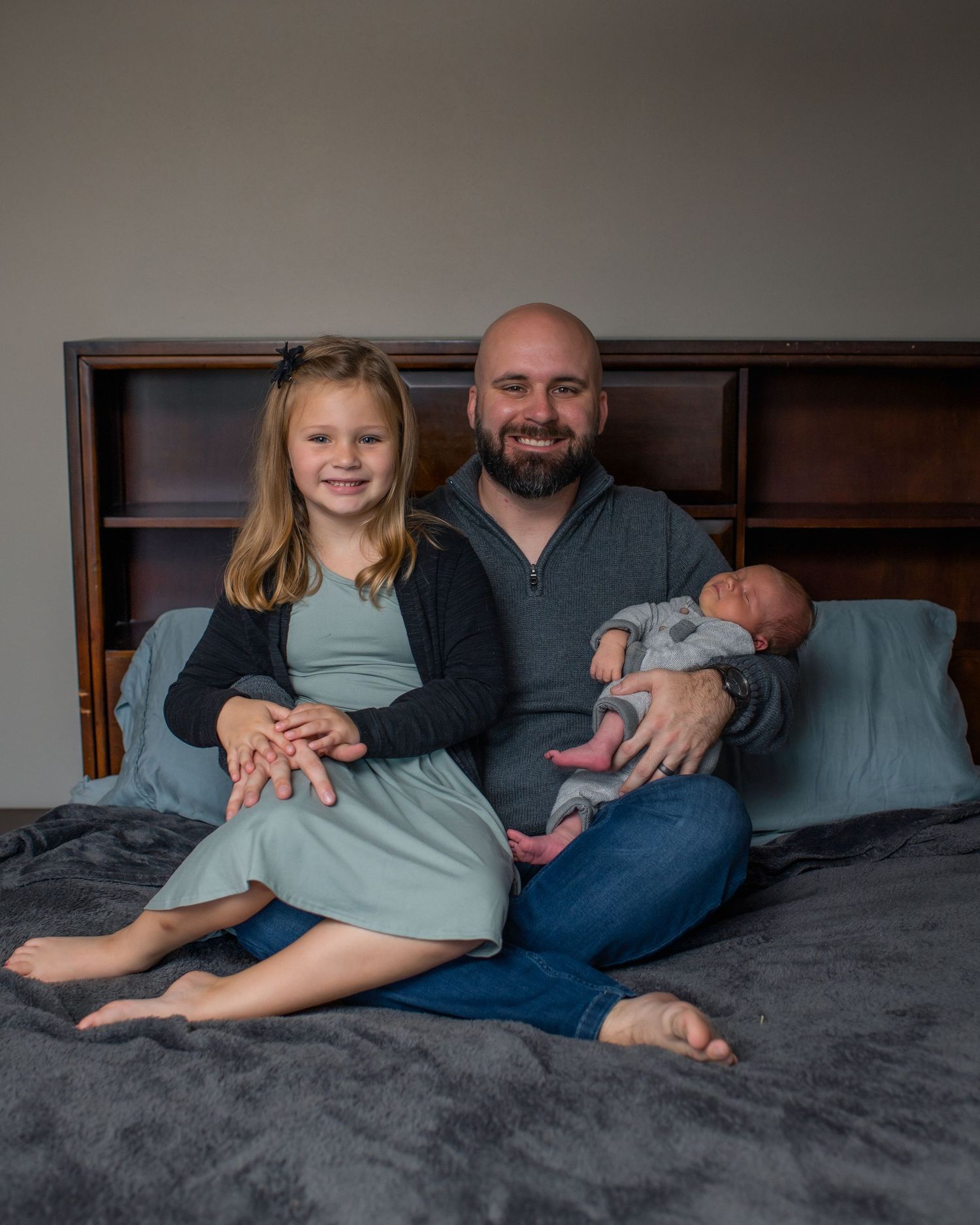 Casual family portrait on bed features father holding newborn while sitting with daughter.