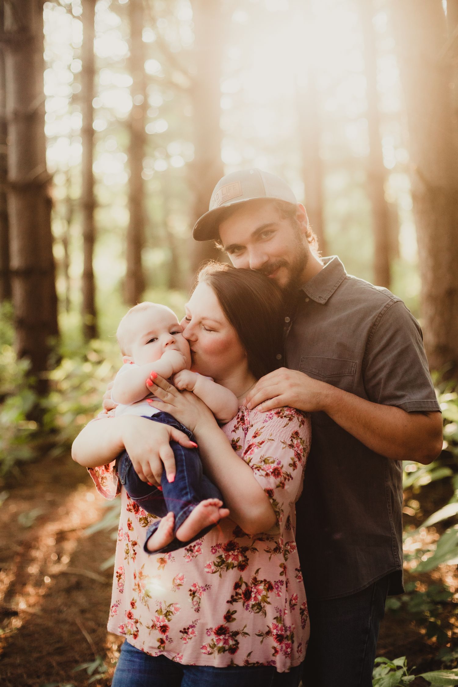two parents stand together in a pine tree forest at sunset; the mother is snuggling baby while dad hugs her from behind