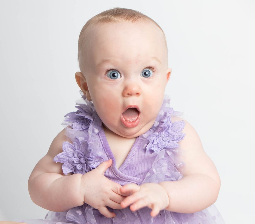 Adorable baby girl in purple ruffle dress with blue eyes and surprised expression in studio.