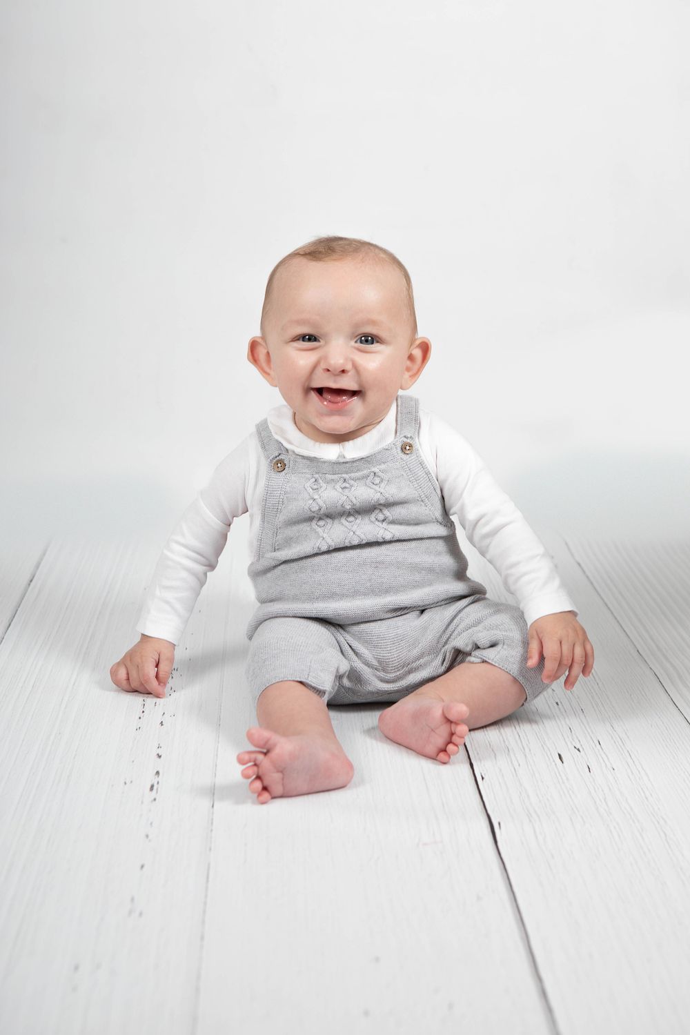 Happy smiling baby boy in grey overalls sitting on white wooden floor in studio portrait.