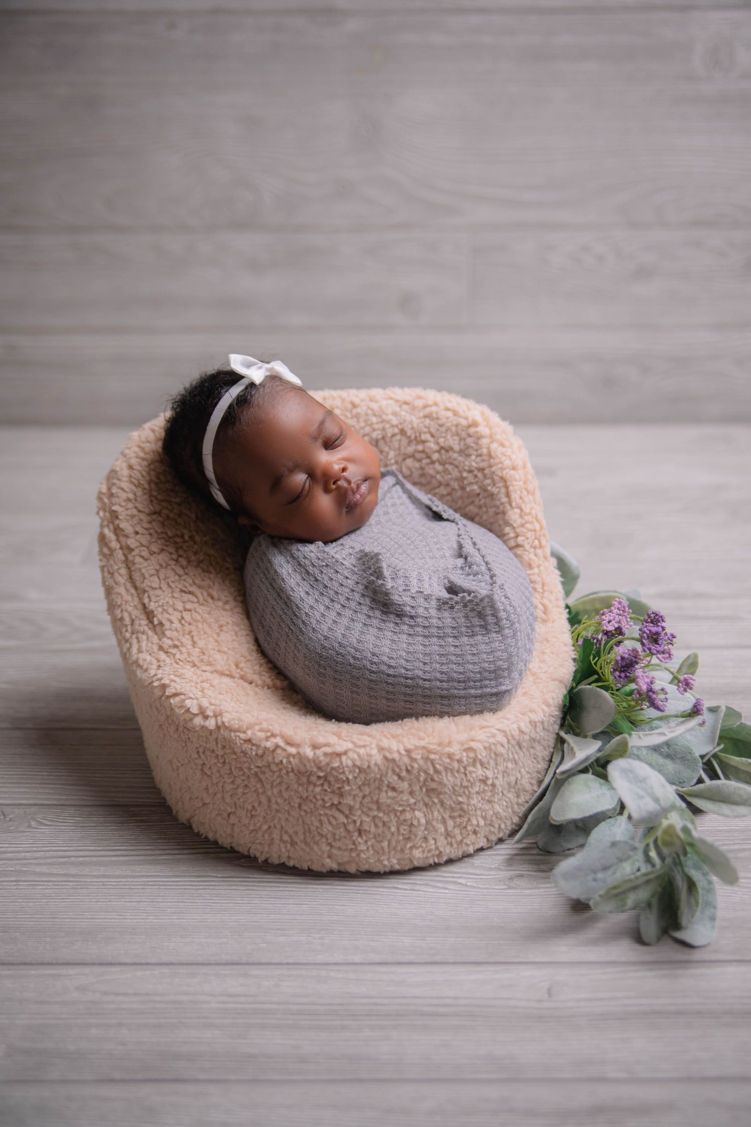 Peaceful newborn portrait on beige round cushion with grey swaddle and eucalyptus decor.