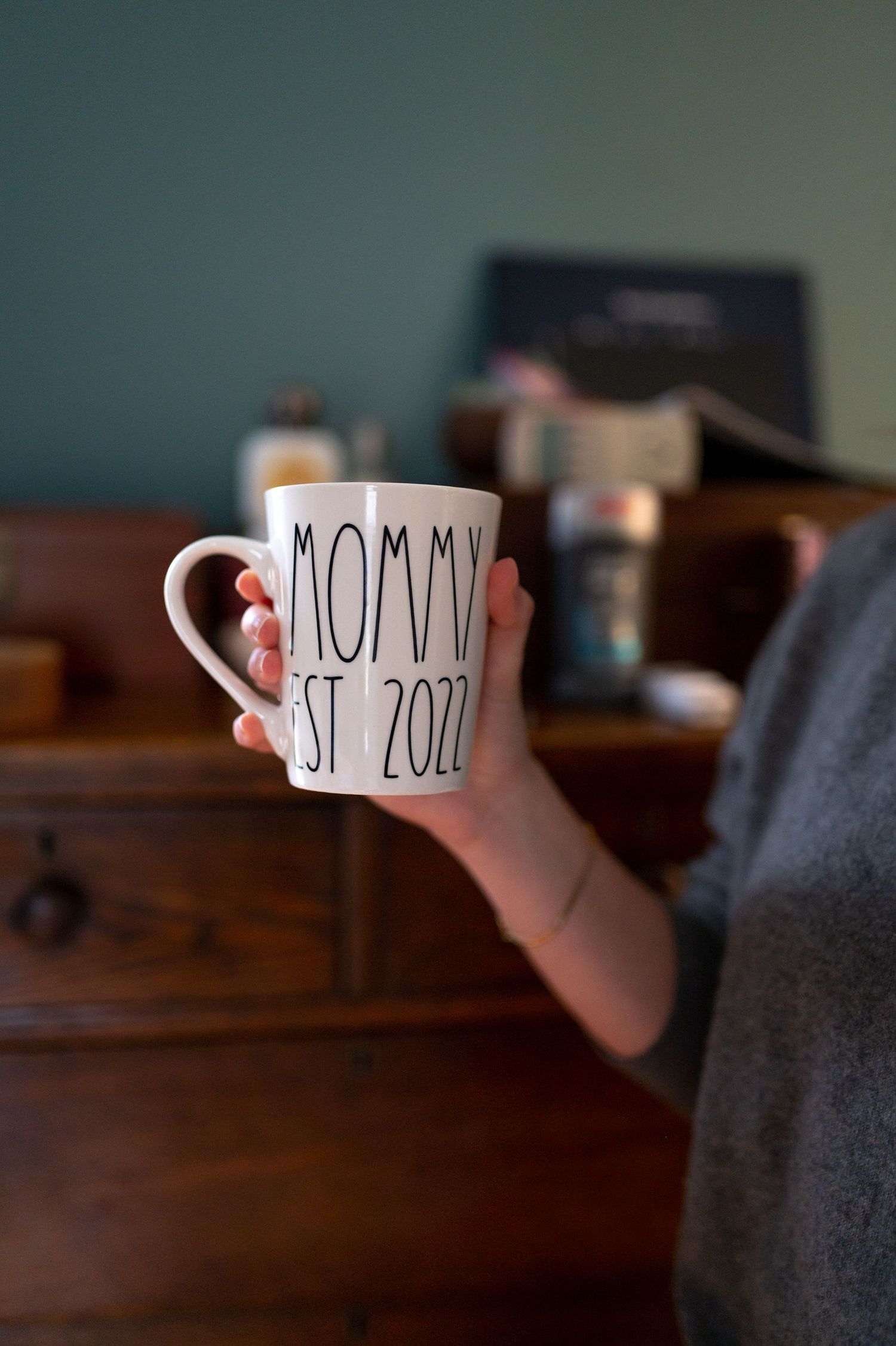 Hand holding a white ceramic mug with 'MOMMY EST 2021' text against a wooden surface.