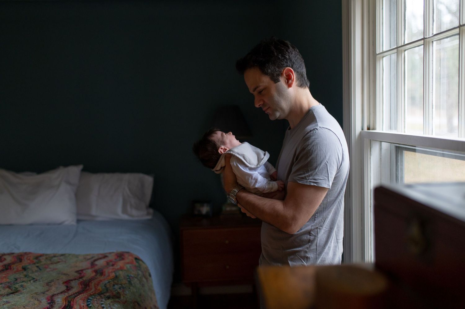 Figure holding newborn baby by window in dimly lit room.