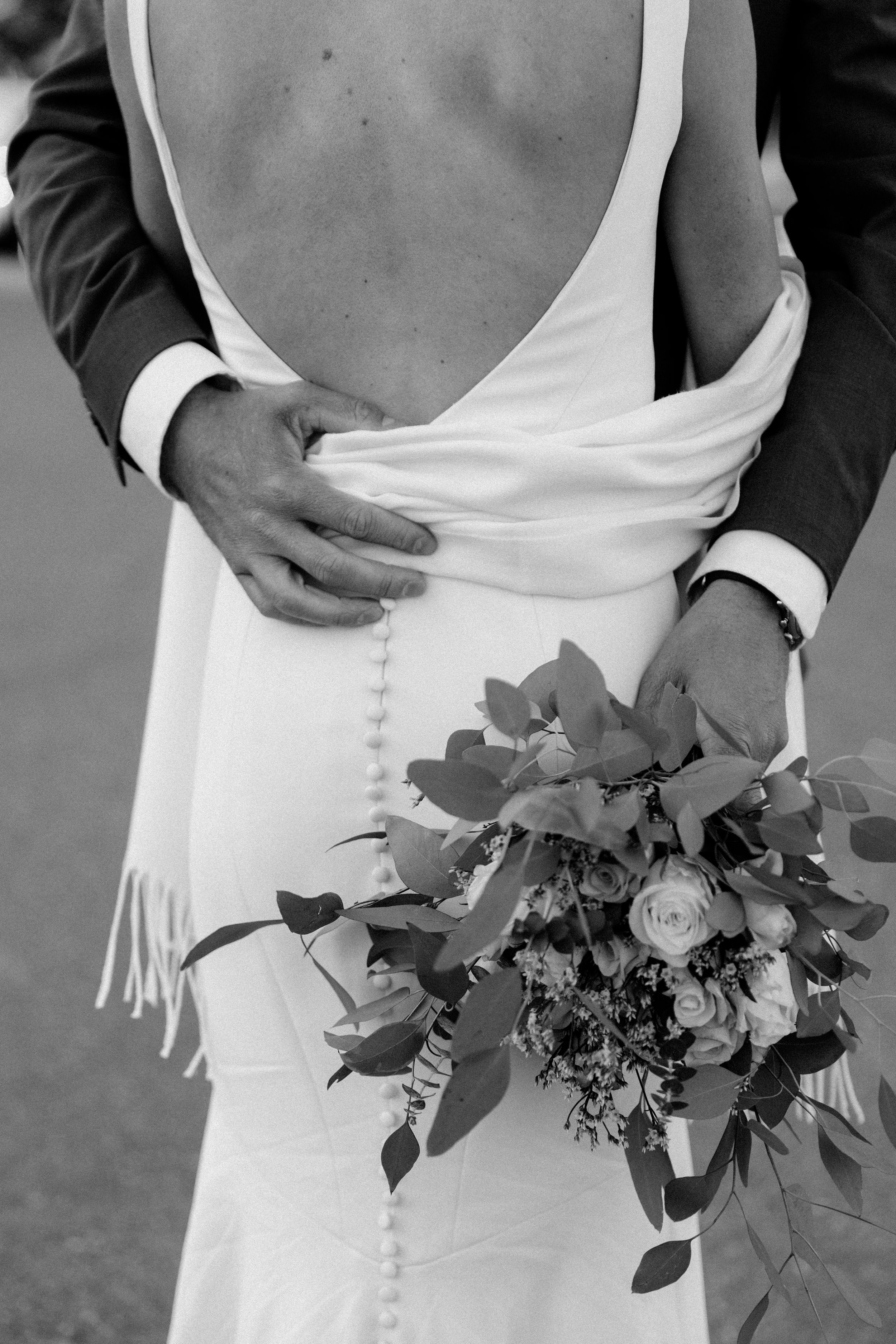 Detail shot of a wedding dress back with buttons and a floral bouquet against a dark suit.
