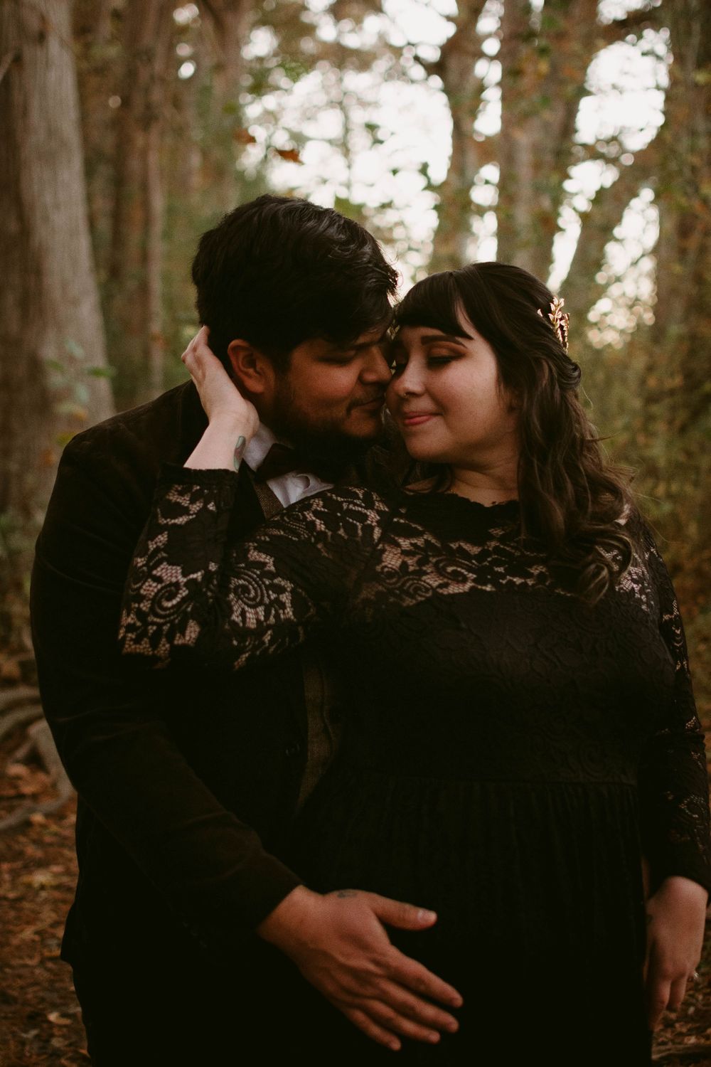 A couple shares an intimate moment wearing matching black outfits in a wooded forest setting.