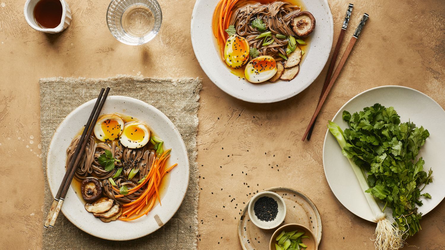 Ramen noodle bowls with soft boiled eggs, vegetables and chopsticks served on textured beige surface.