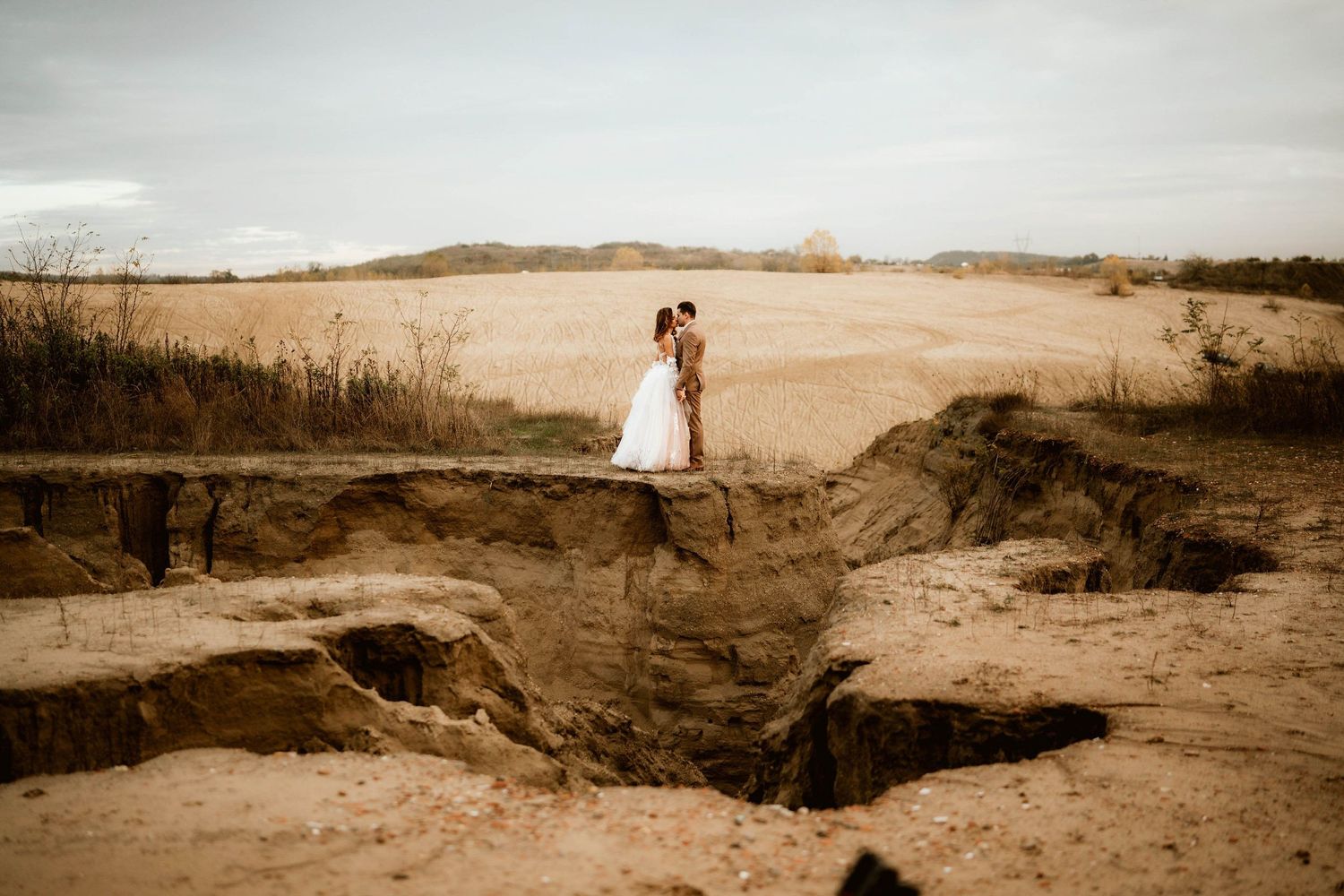 Romantic wedding photo of couple standing on sandy cliff edge at sunset.