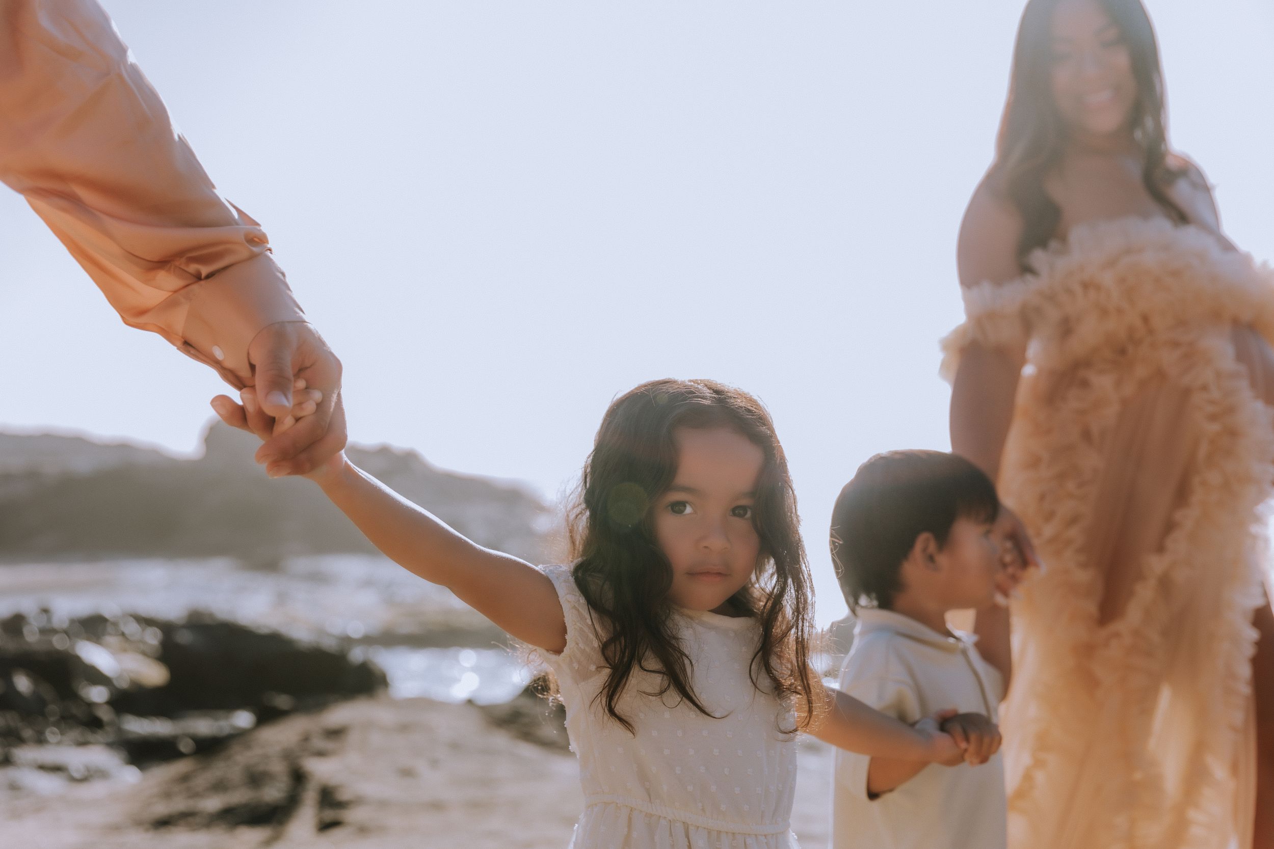 People in light flowy outfits gather on beach during golden hour.