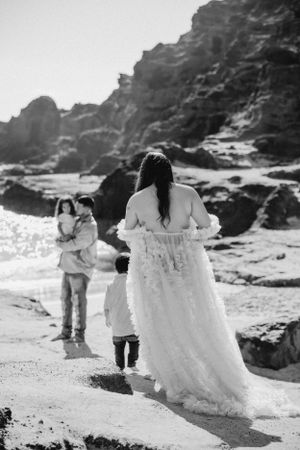 Beach maternity session with white dress against rocky coastal backdrop.