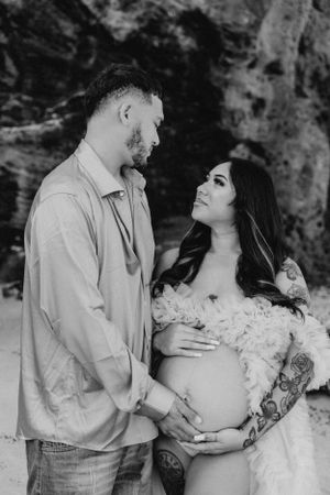 A romantic maternity photoshoot on a beach with a couple embracing in front of rocky cliffs.