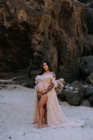 Expectant mother in a flowing peach gown poses gracefully against rocky coastal cliffs.