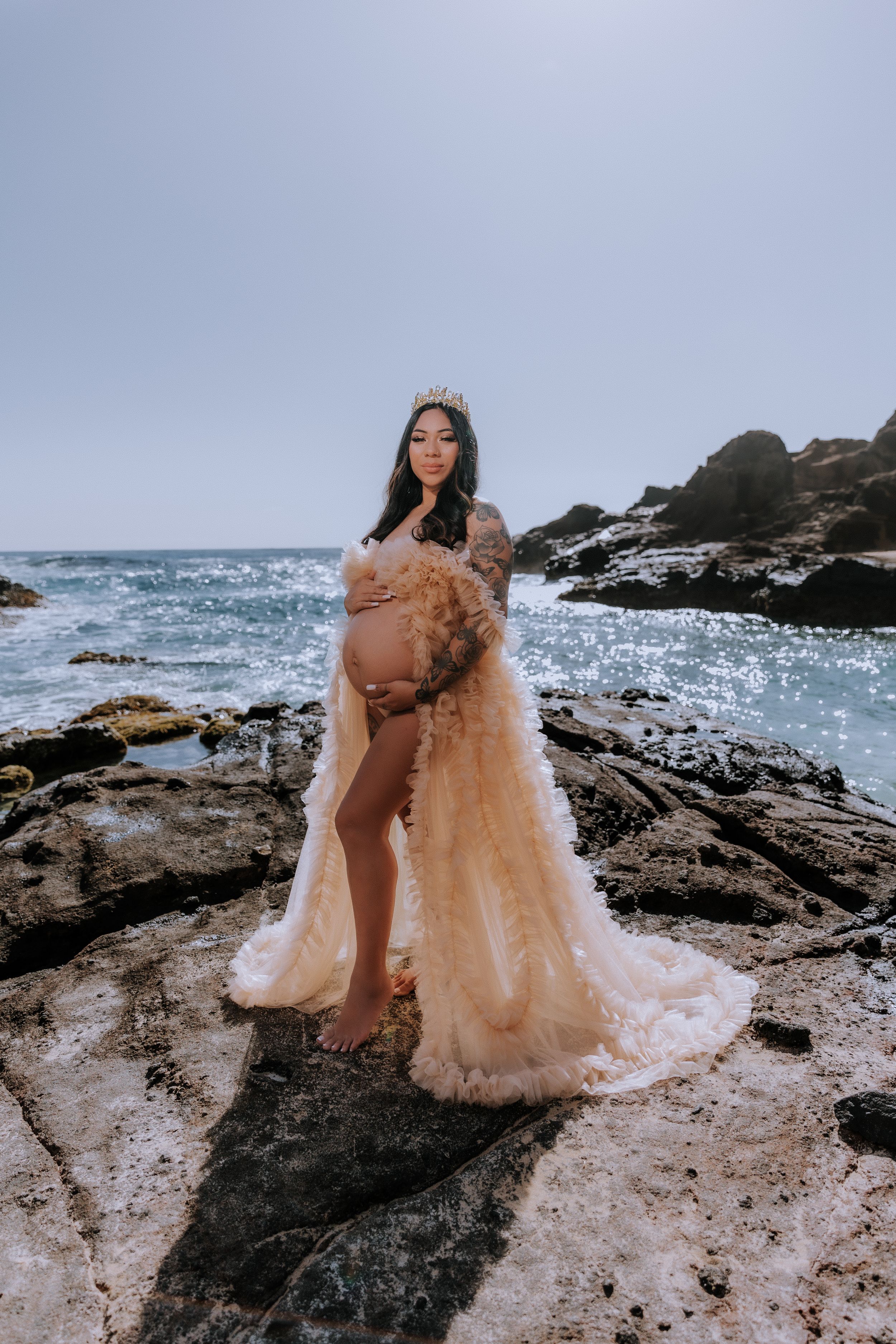 Peachy tulle maternity gown flowing in ocean breeze against rocky coastline.
