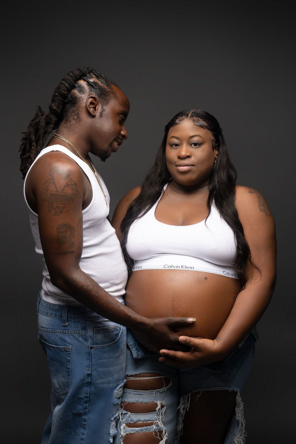 Intimate maternity photoshoot against dark background with couple in white tops and distressed jeans.