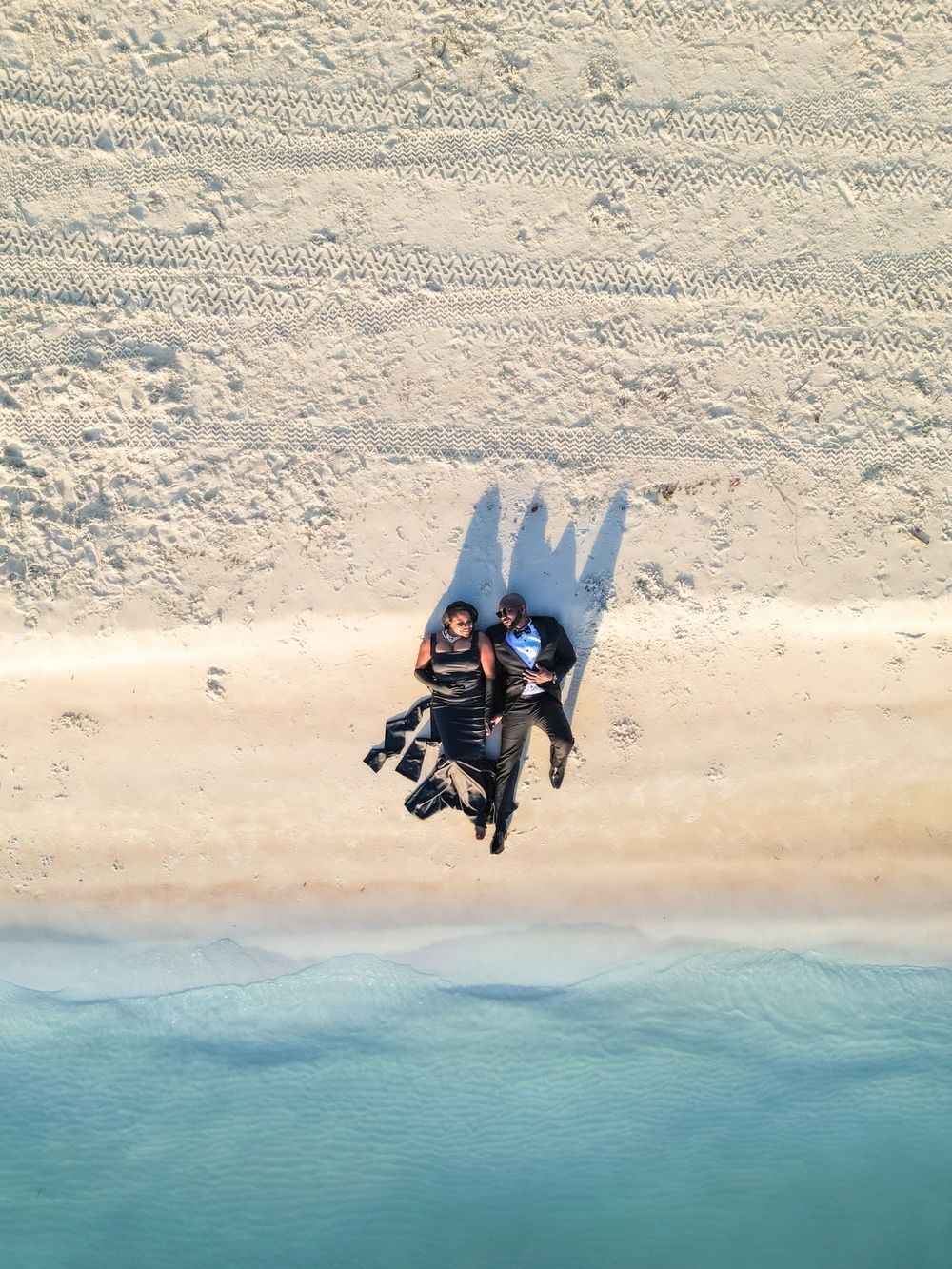 Aerial view of two people lying on sandy beach with long shadows cast on the sand.