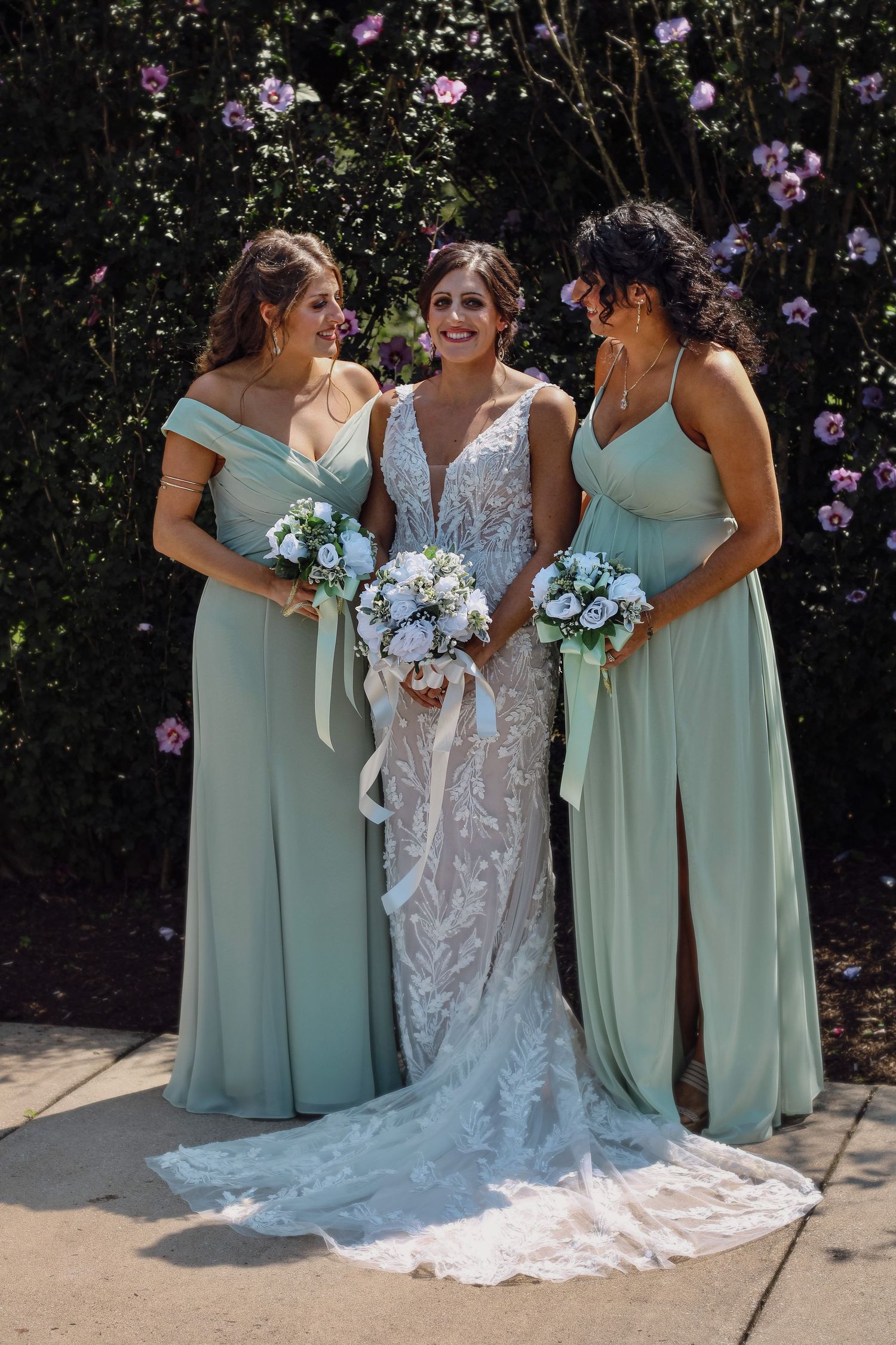 Three women in mint green bridesmaids dresses and white lace wedding gown pose together against floral backdrop.