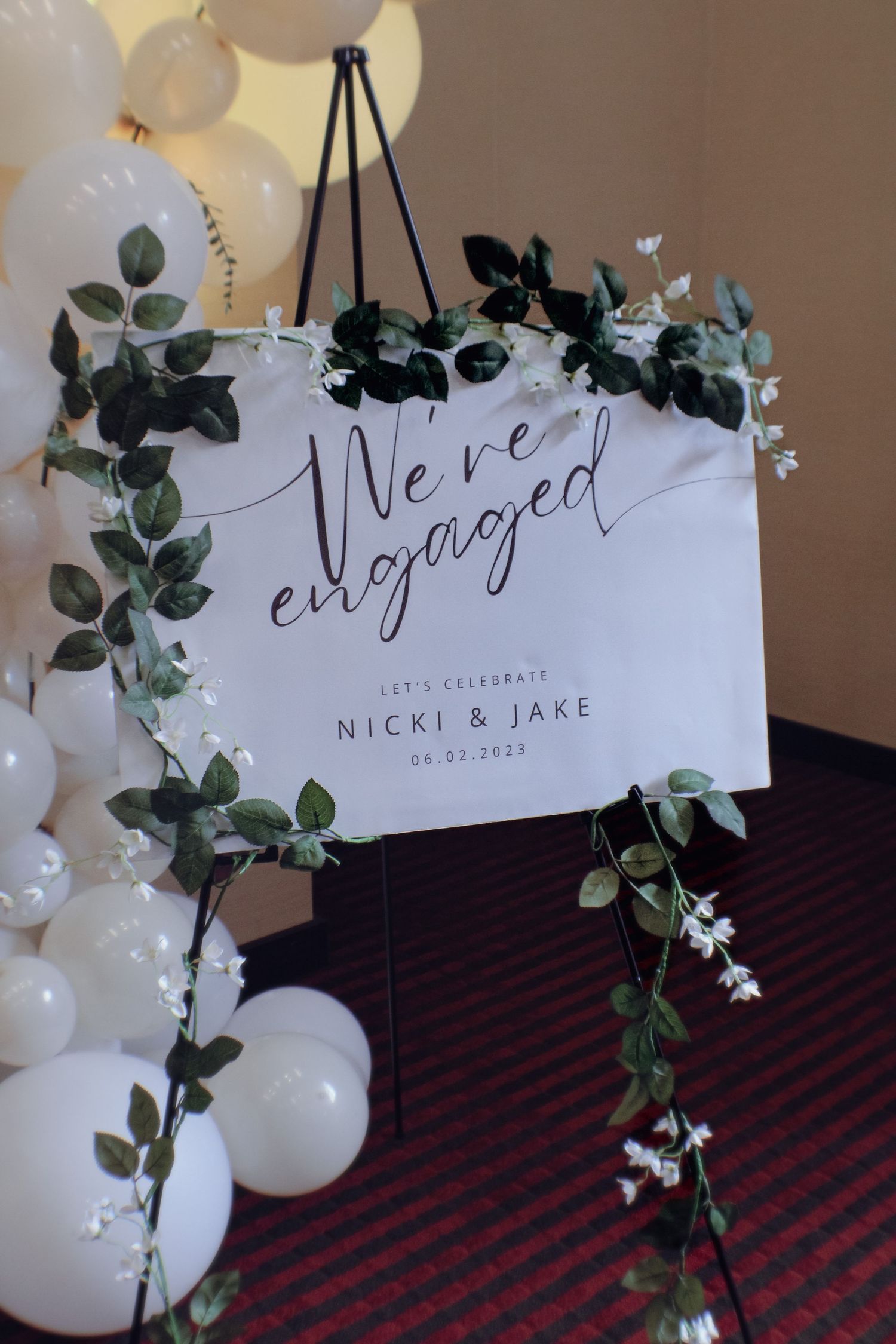 Elegant engagement announcement sign with 'We're engaged' text decorated with eucalyptus leaves and white balloons.