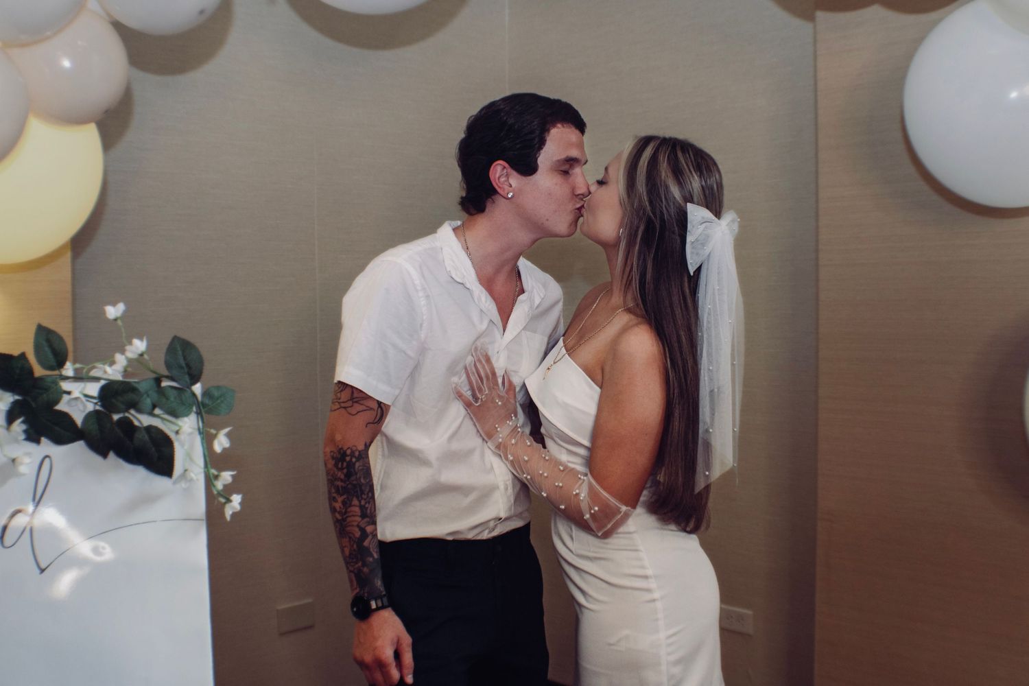 A romantic couple kisses at a wedding celebration with white balloons in the background.