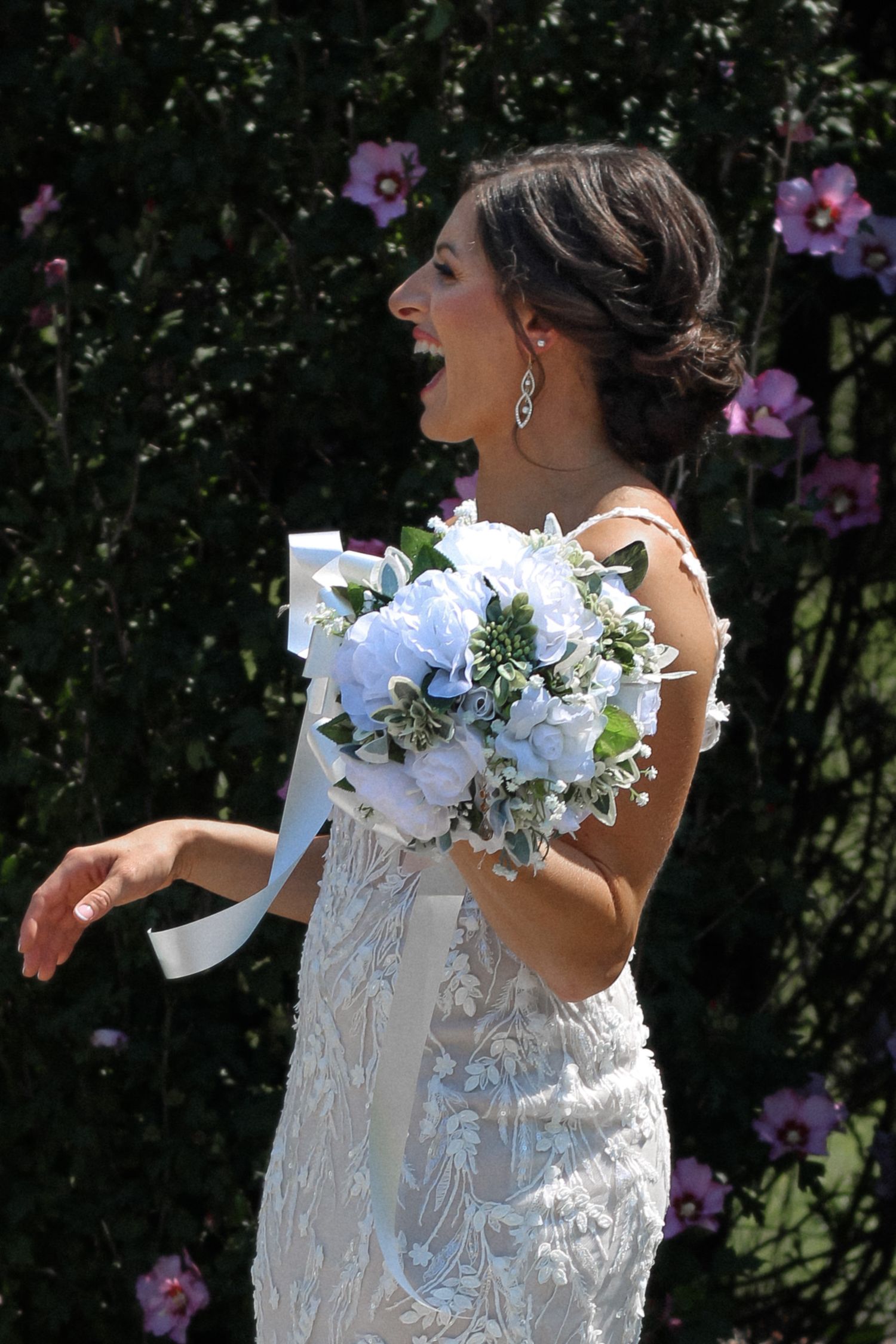 Happy bride in lace gown holds blue and white bouquet against purple flower backdrop.