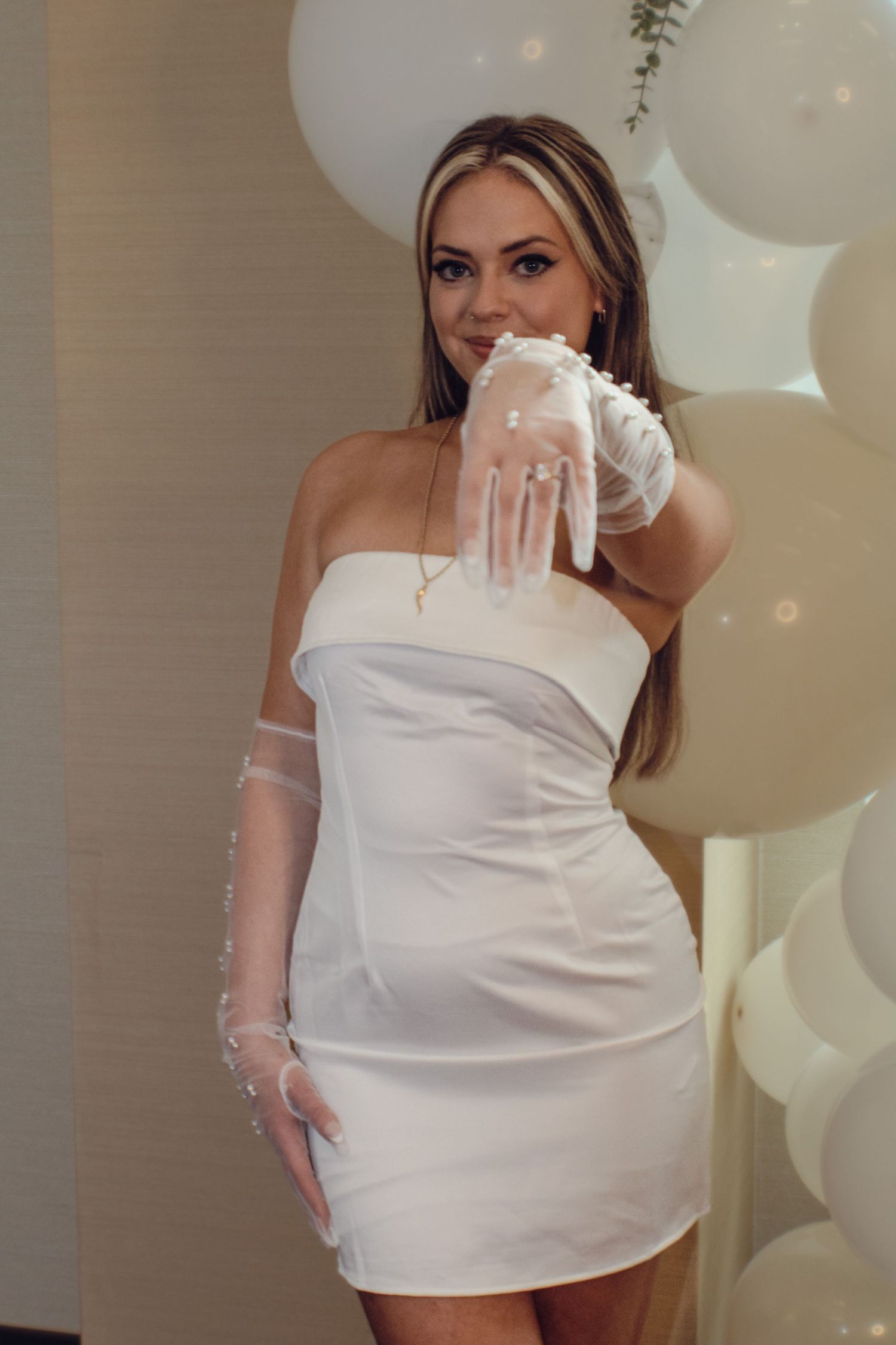 A bride in a short white dress and lace gloves poses against a backdrop of white balloons.