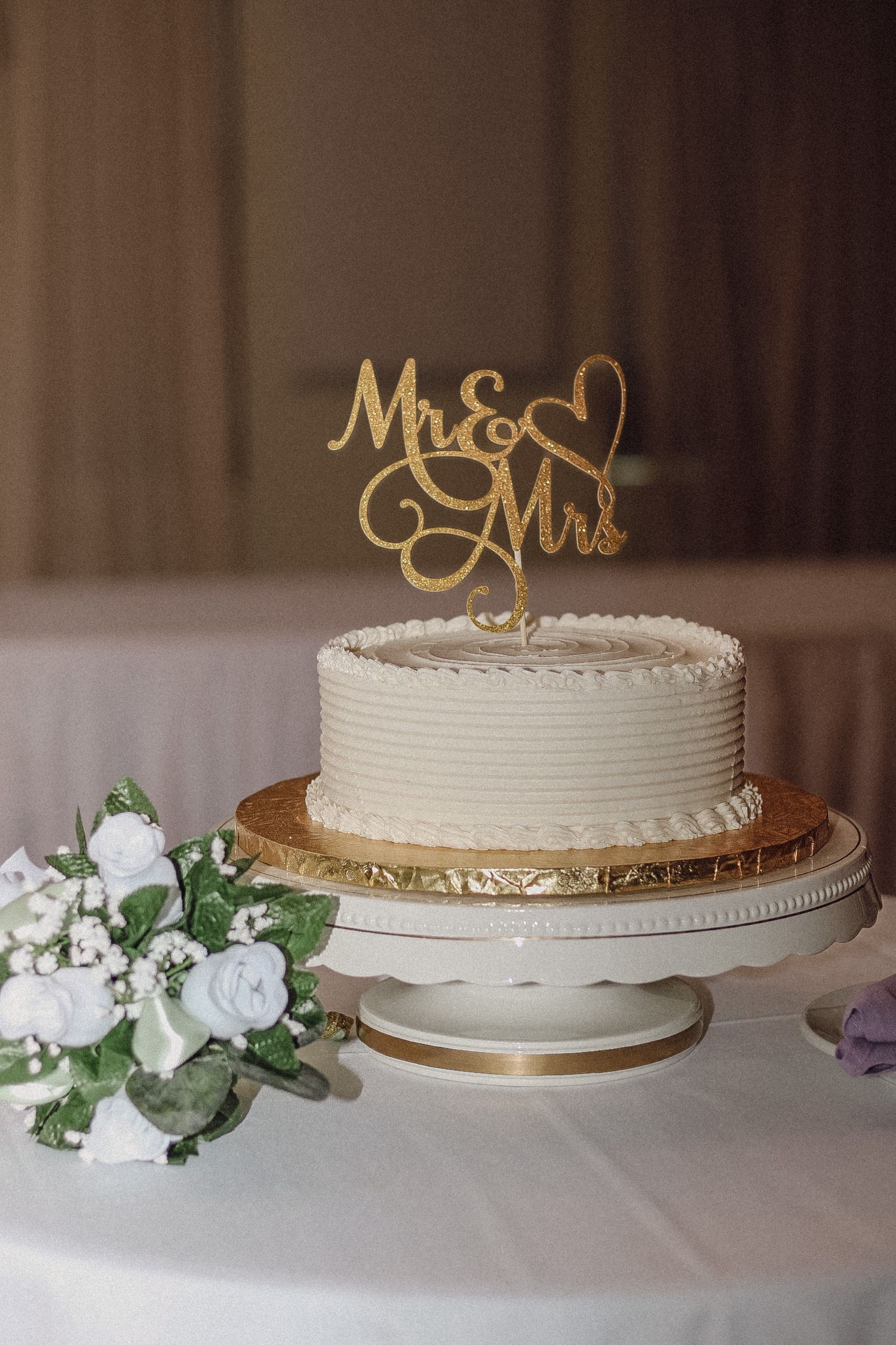 Simple white wedding cake with gold 'Mr & Mrs' topper and floral decorations on gold cake stand.