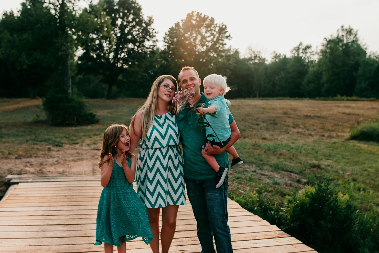 A small family stands together on a wooden boardwalk in a natural outdoor setting.