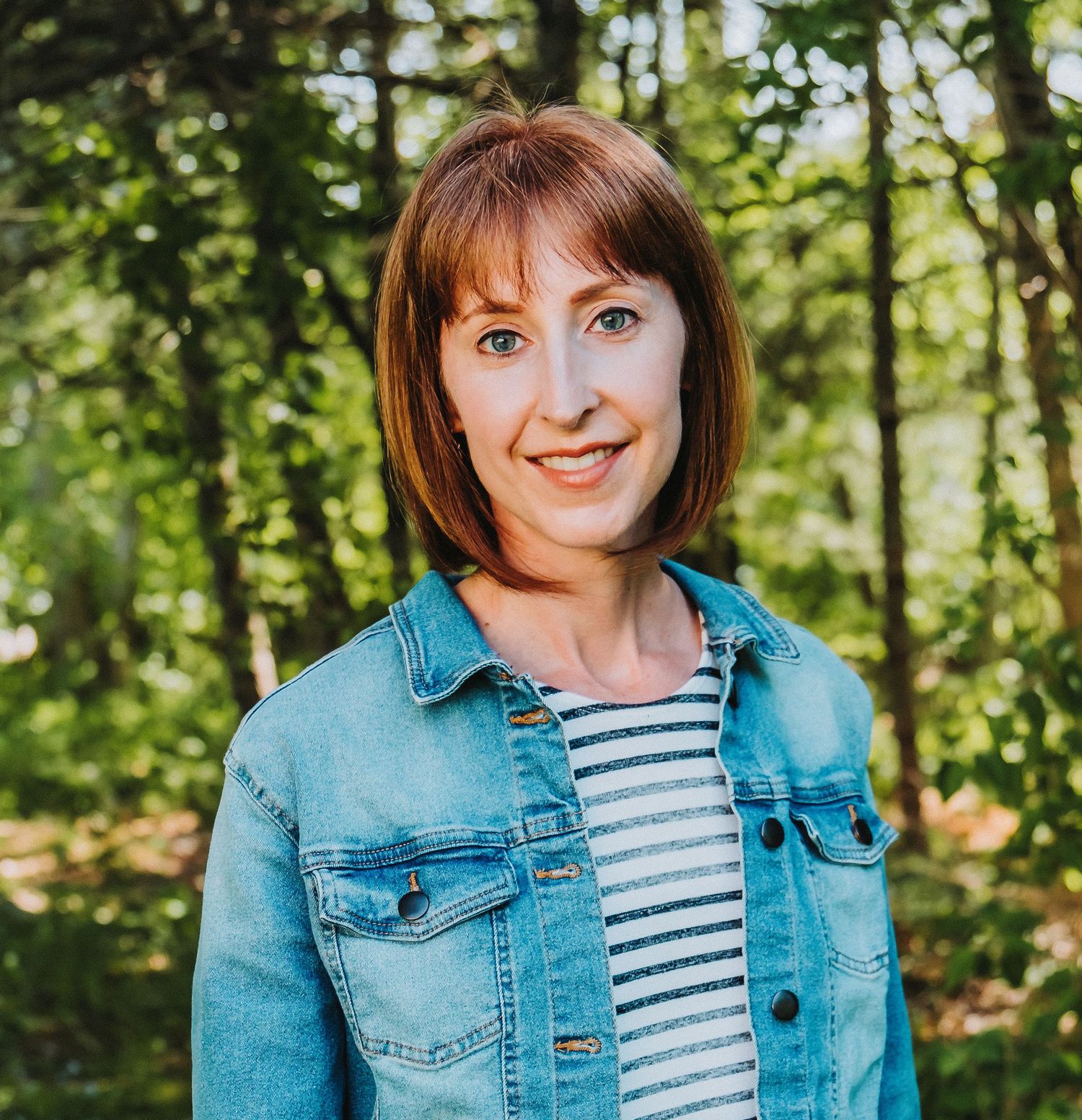 Portrait of someone wearing denim jacket and striped shirt against green forest background.