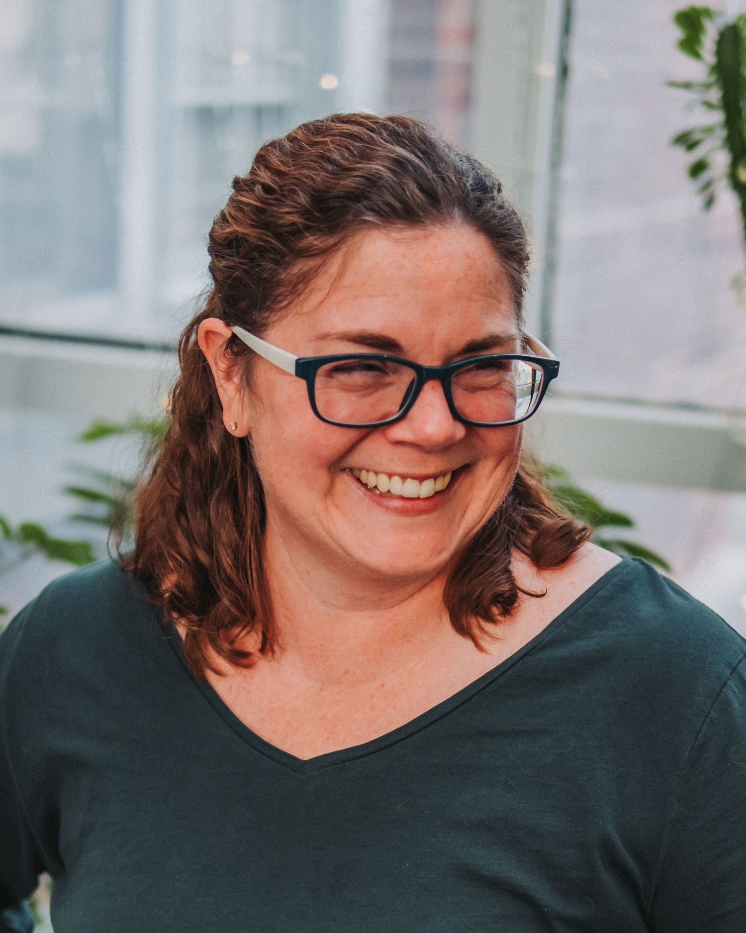 Portrait of person in glasses and dark top smiling warmly against bright window background.