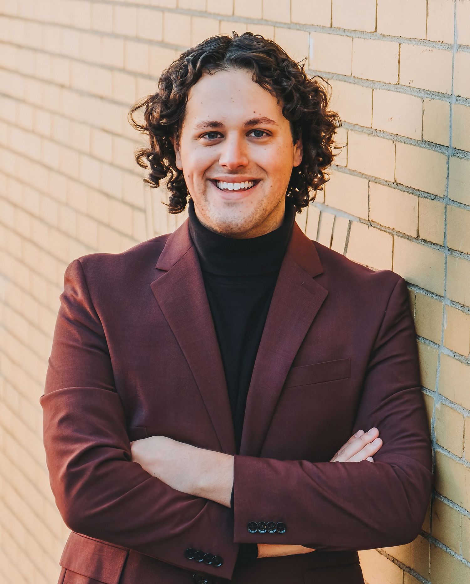 Professional portrait of person in burgundy blazer and black turtleneck against brick wall.