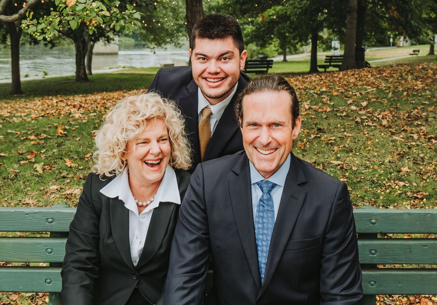 Three business professionals pose together on a park bench surrounded by fall leaves.
