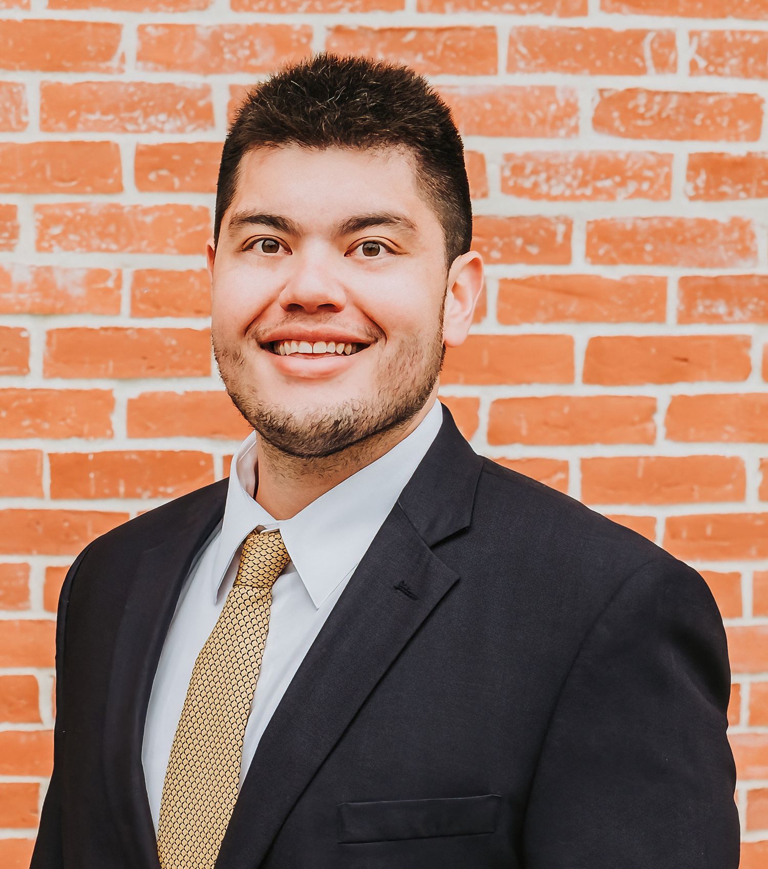 Professional headshot of person in dark suit with gold tie against orange brick wall.