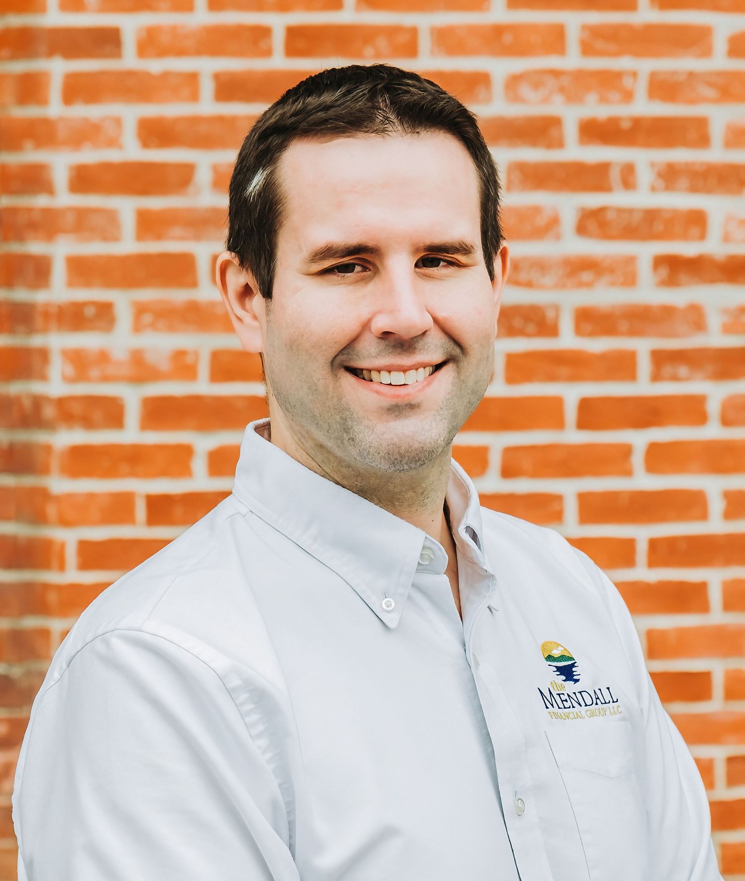 Corporate headshot of person in white button-down shirt with company logo against brick wall.