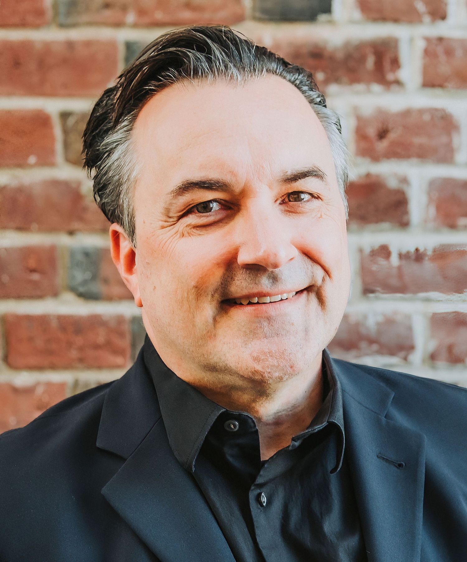 Professional portrait of a person in a black suit jacket against a brick wall background.