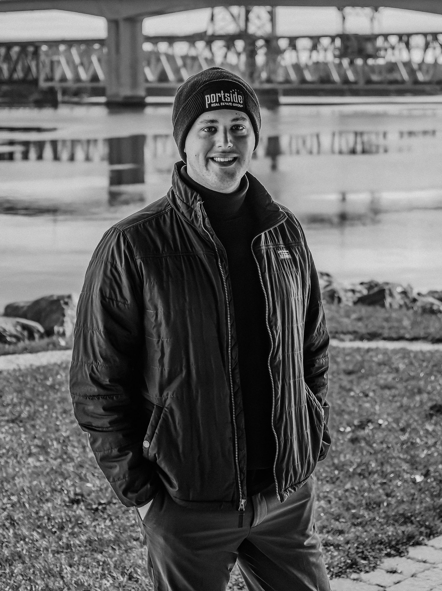 Black and white portrait of person in winter jacket and beanie smiling by riverside.