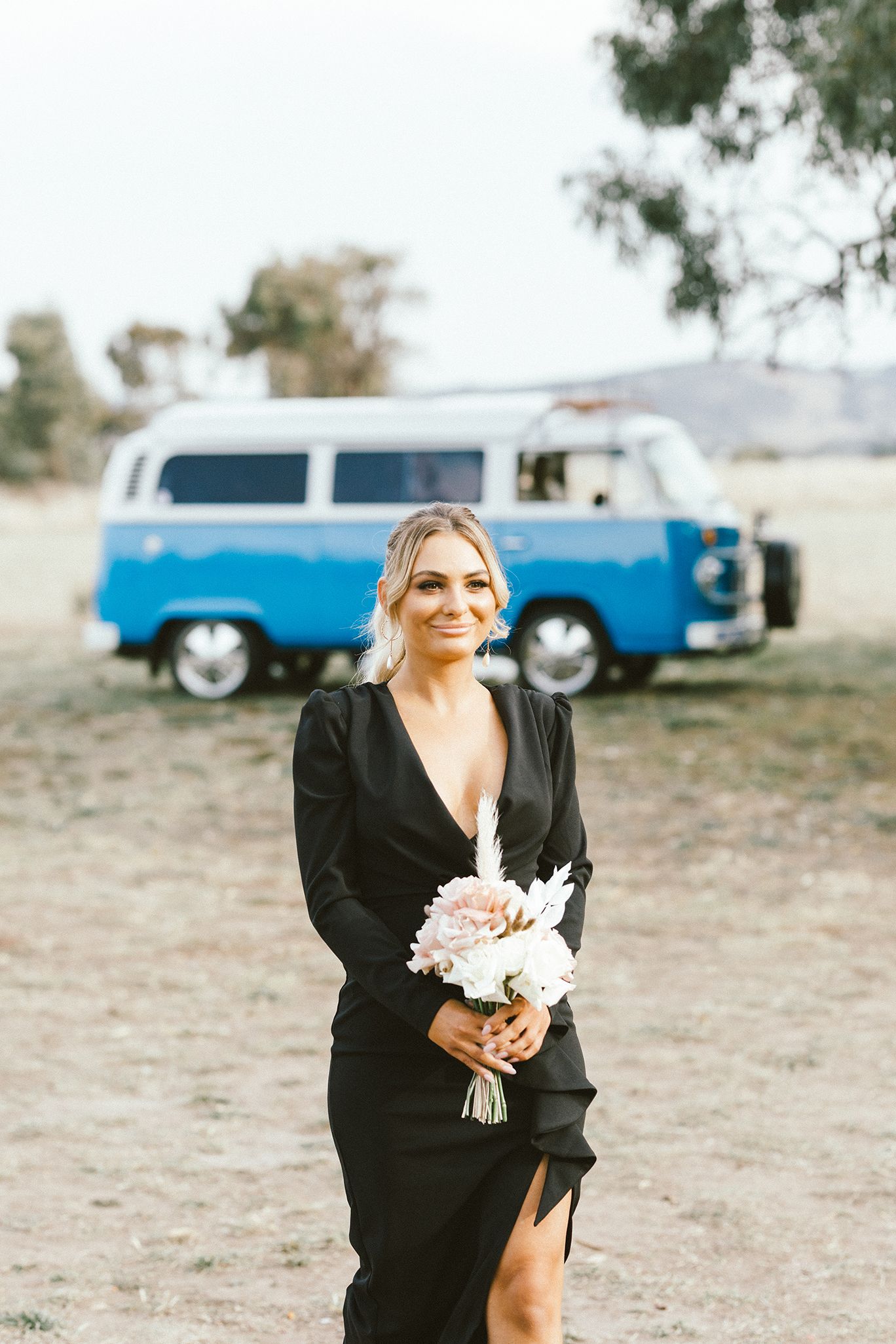 Elegant bridesmaid in black dress holding bouquet with classic blue VW van behind.