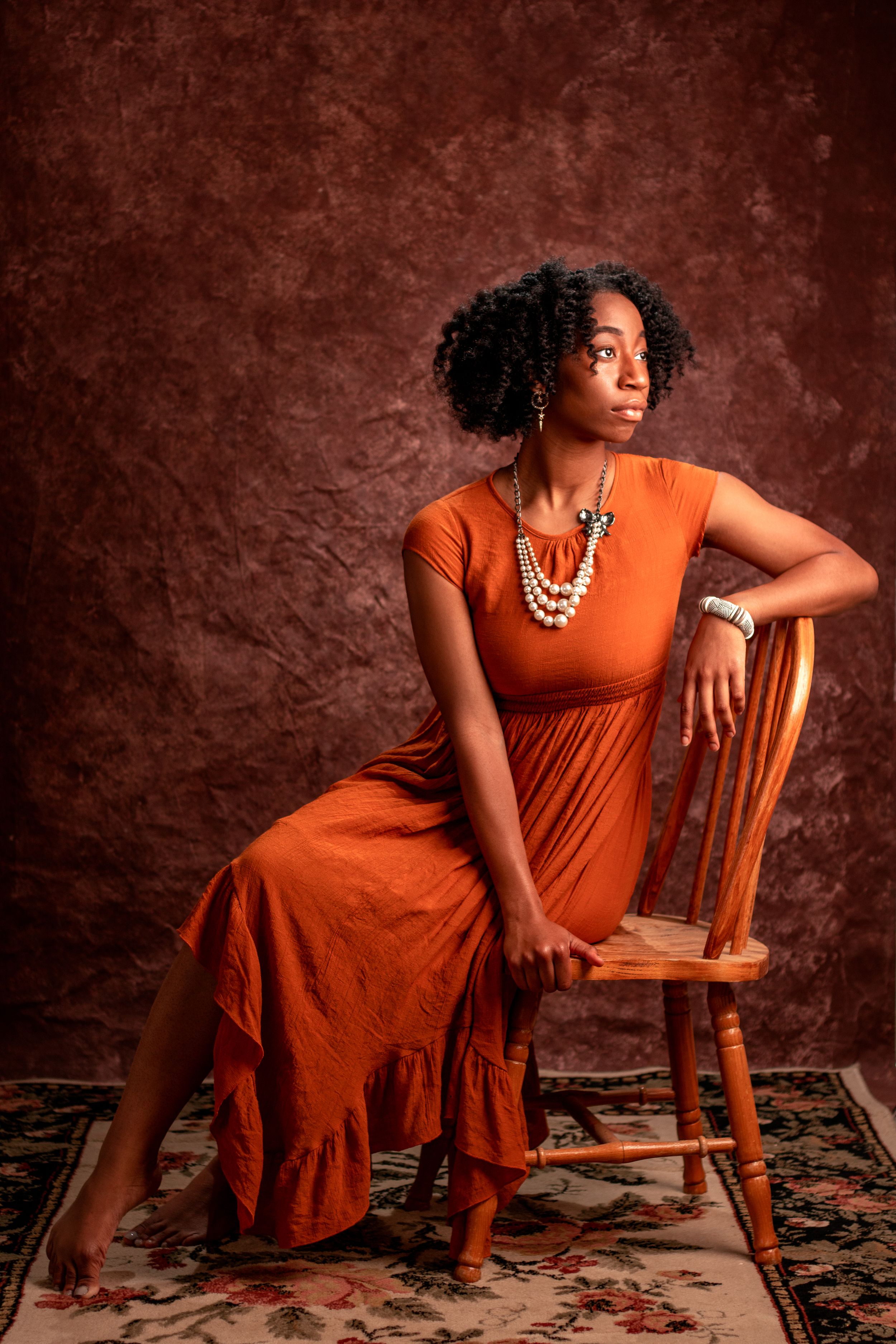 A portrait session featuring someone in an orange dress posed elegantly on a wooden chair.